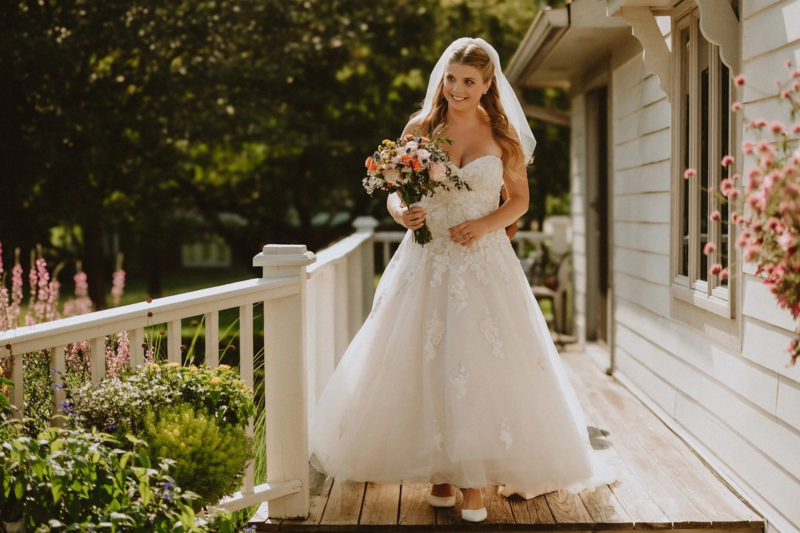 Bride in white wedding dress holding bouquet on porch with flowers and railing.