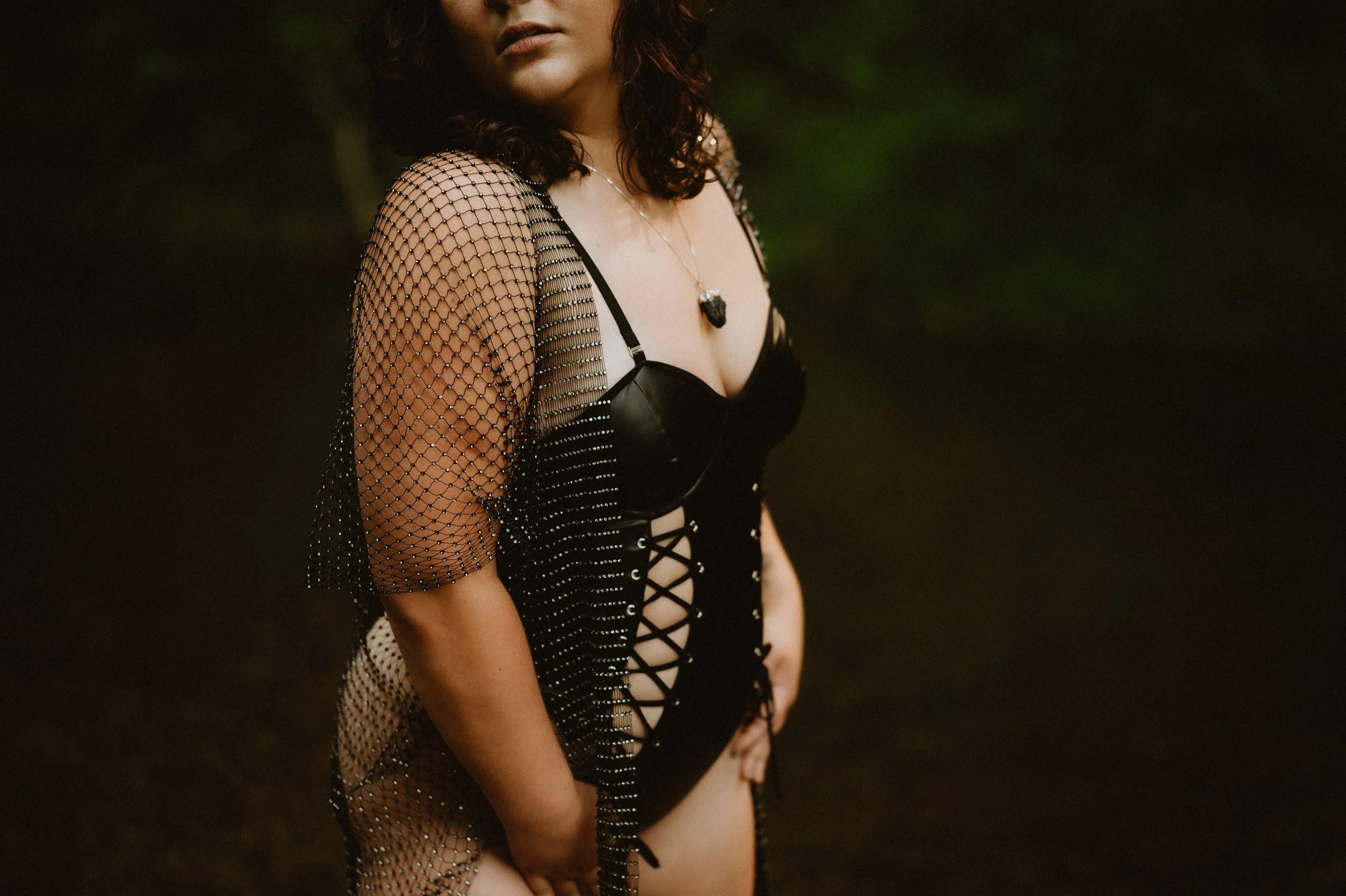 Woman in black lace-up bodysuit and netted cover-up, wearing a necklace, standing outdoors.