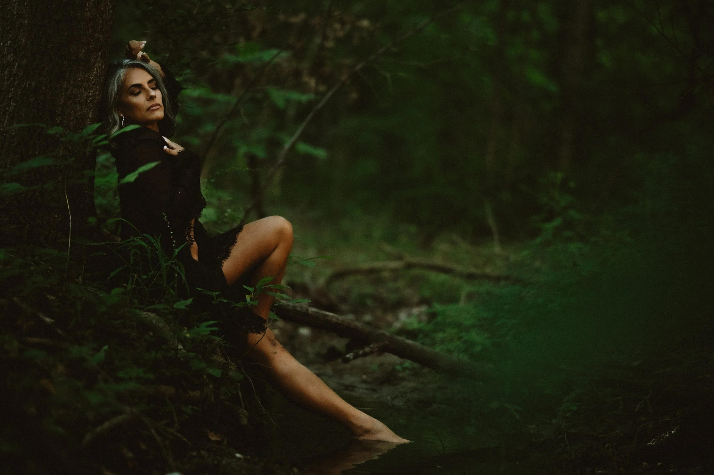 Woman in dark clothing sitting against a tree in a forest setting, legs partially submerged in water.