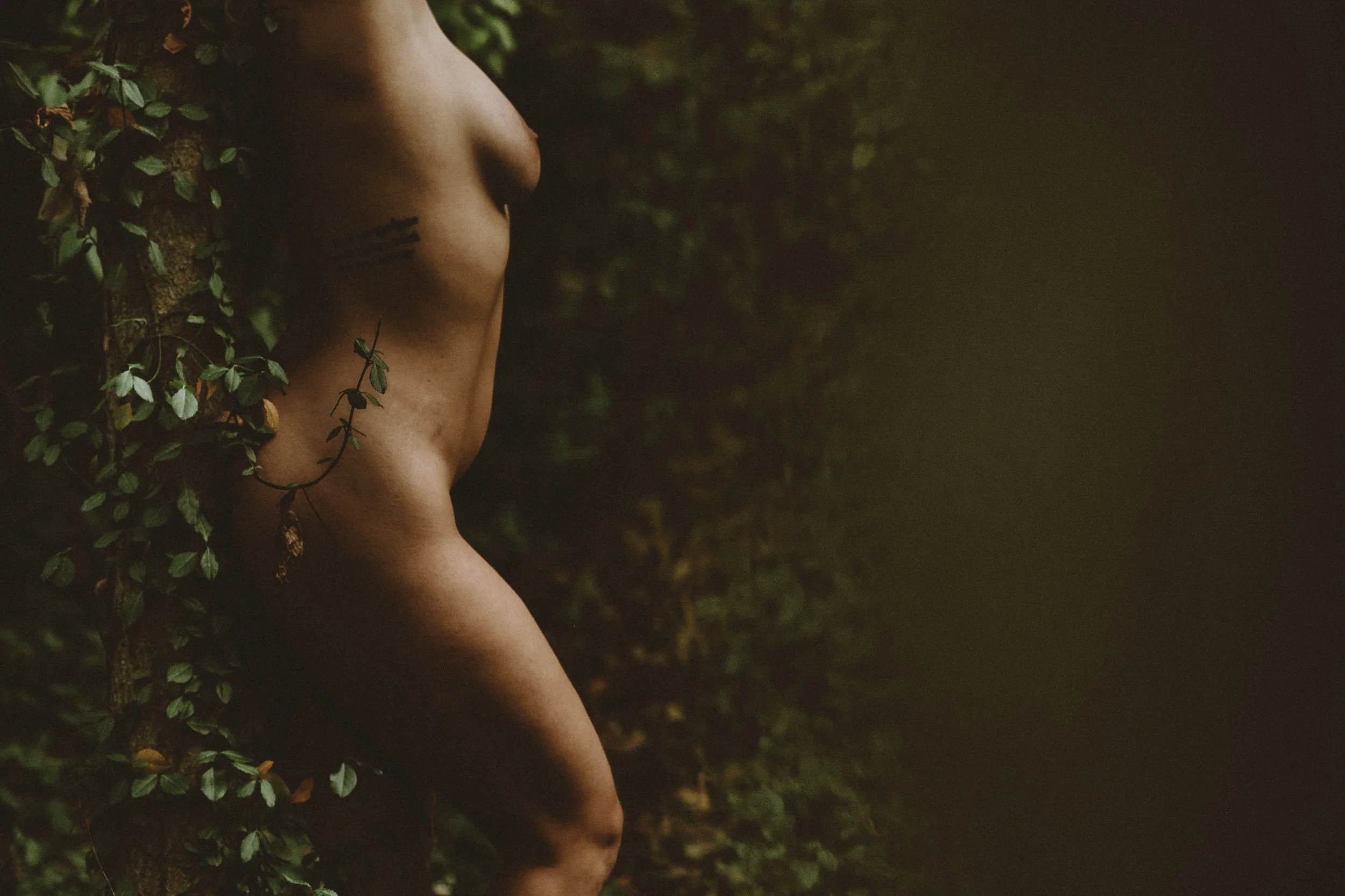 Partial view of a person standing in nature, with vines and leaves, showcasing a tattoo on the side.