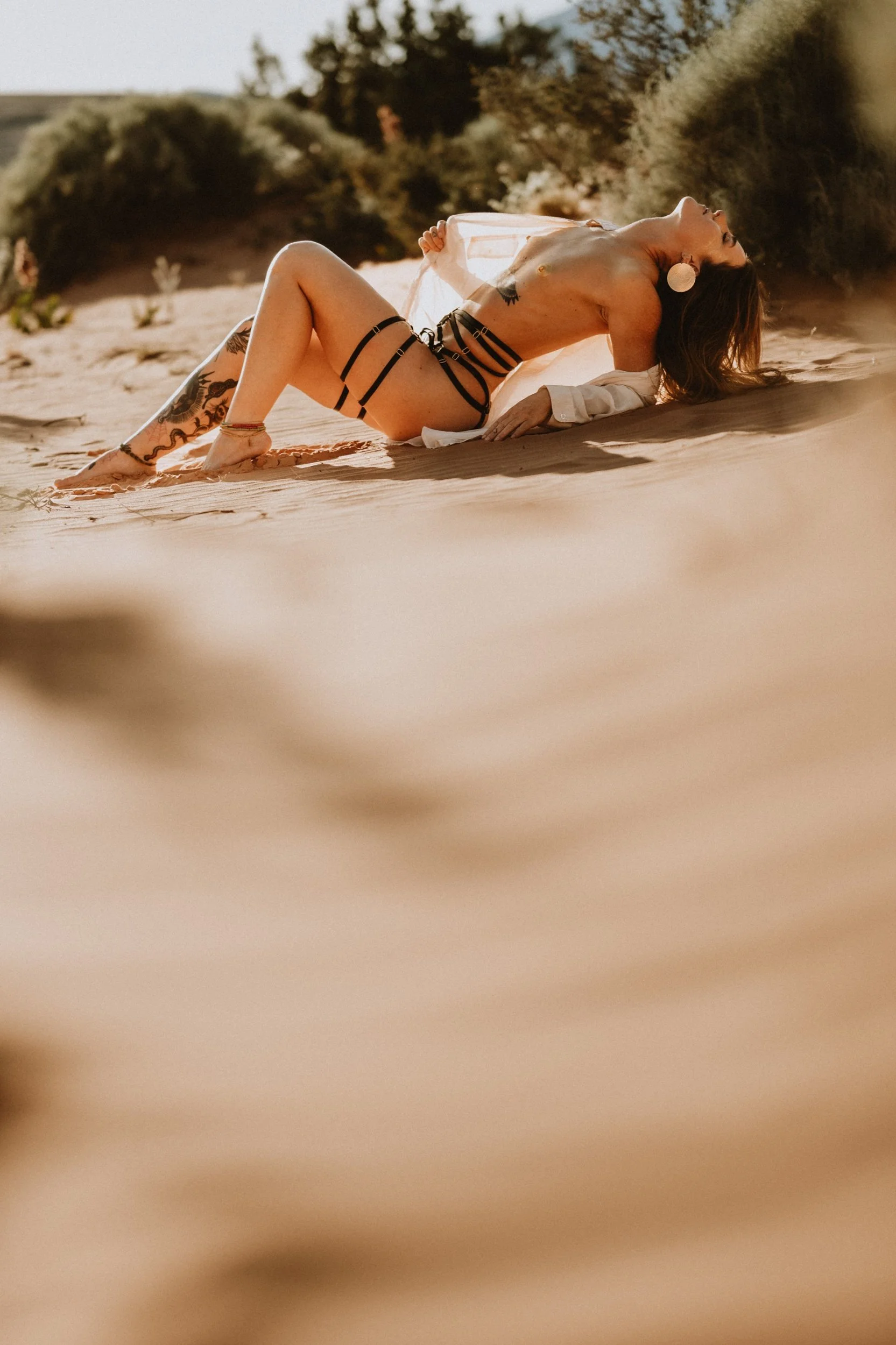 Person in black strappy swimsuit poses on sandy beach with greenery in background.