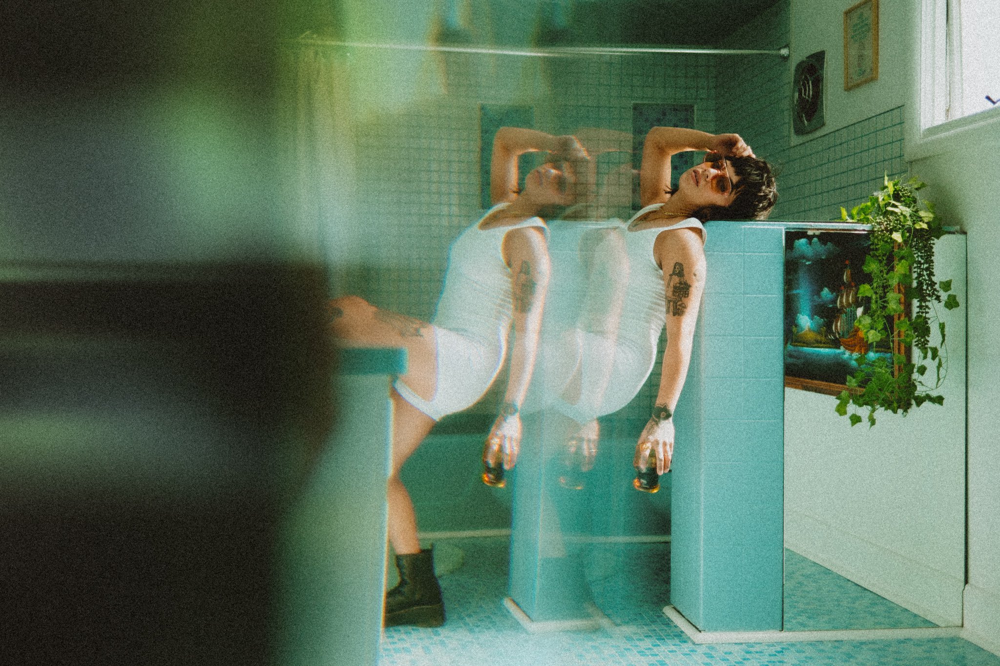 Person wearing a white tank top and shorts, sitting on a bathroom counter with a blurred effect. They have tattoos, sunglasses, and are holding a drink, with a plant and framed picture in the background.