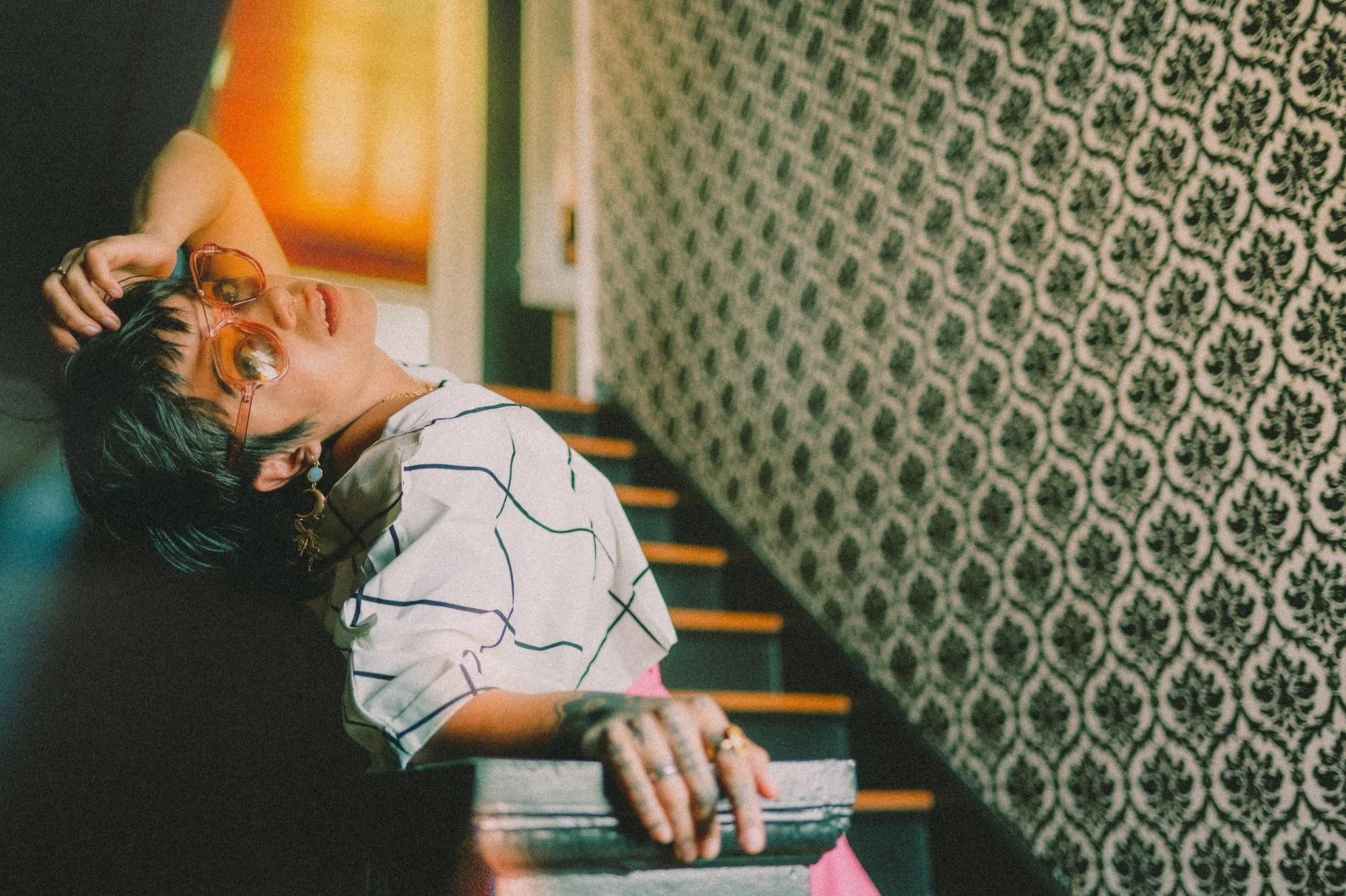 Person wearing sunglasses and patterned shirt leaning on staircase railing, with patterned wallpaper background.