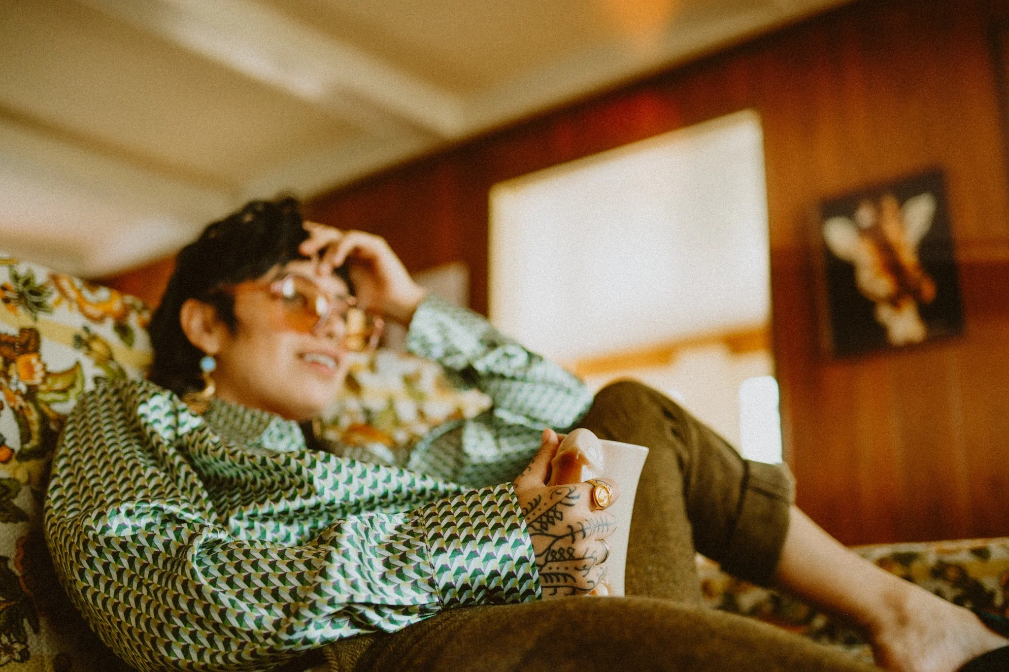 Person relaxing on a floral couch, wearing sunglasses and a patterned shirt, holding a white mug, with tattoos on their hand.