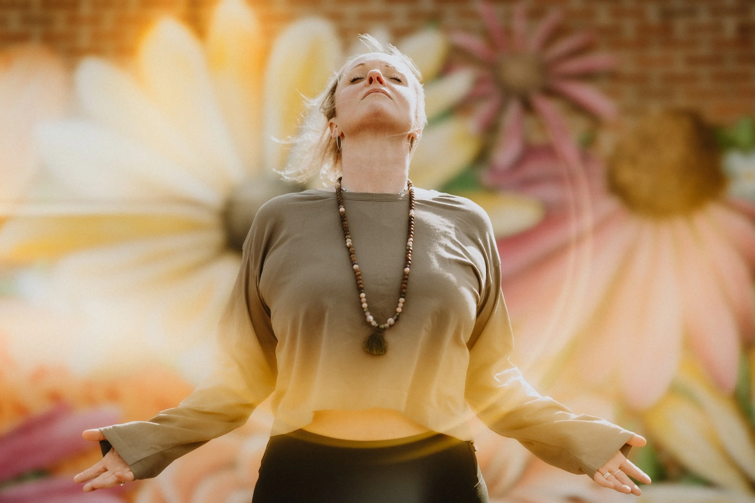 Woman in brown shirt practicing yoga outdoors against a floral mural background.