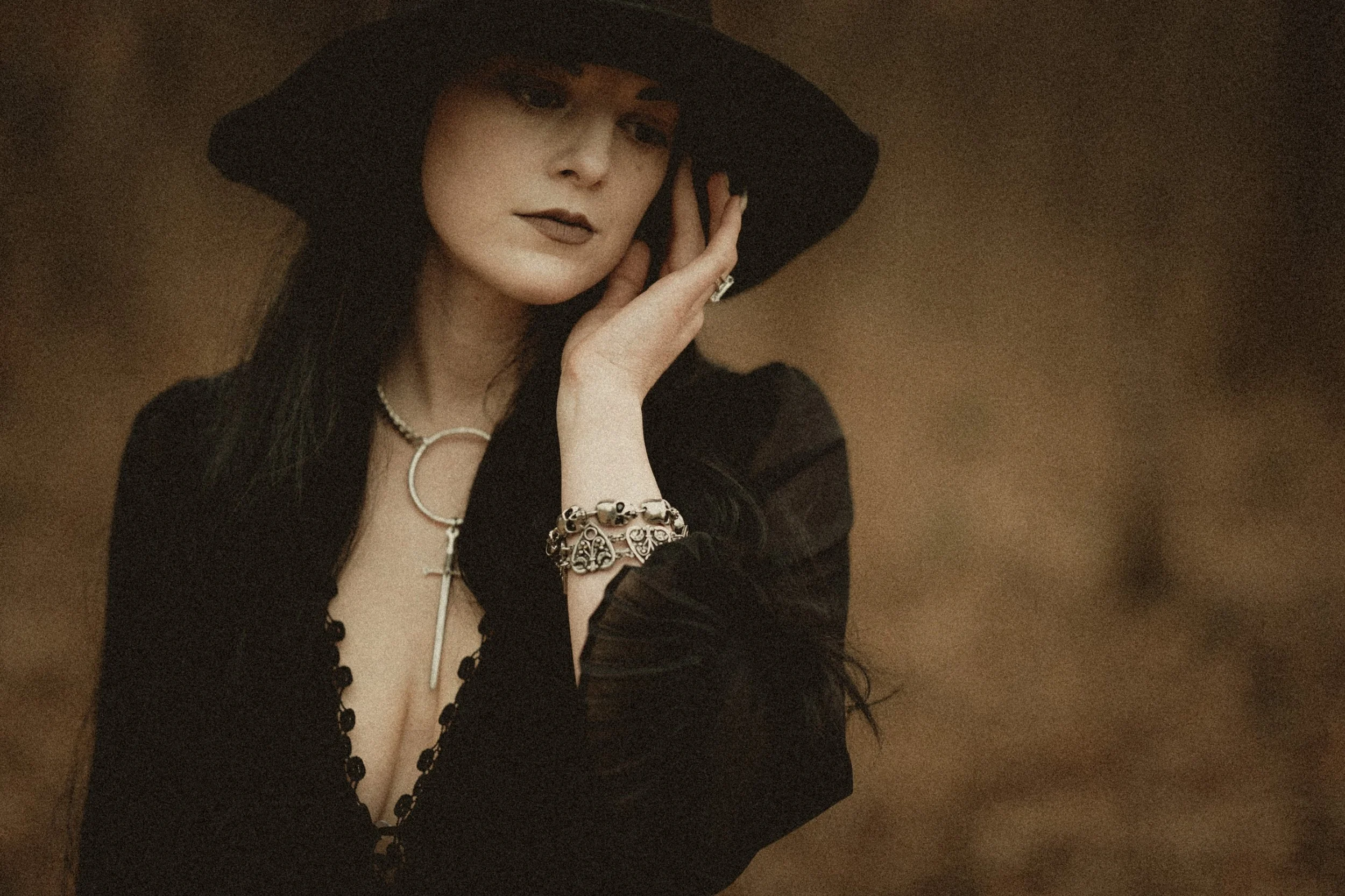 Woman in dark clothing with a wide-brimmed hat, wearing a cross necklace and skull bracelet, in a contemplative pose.
