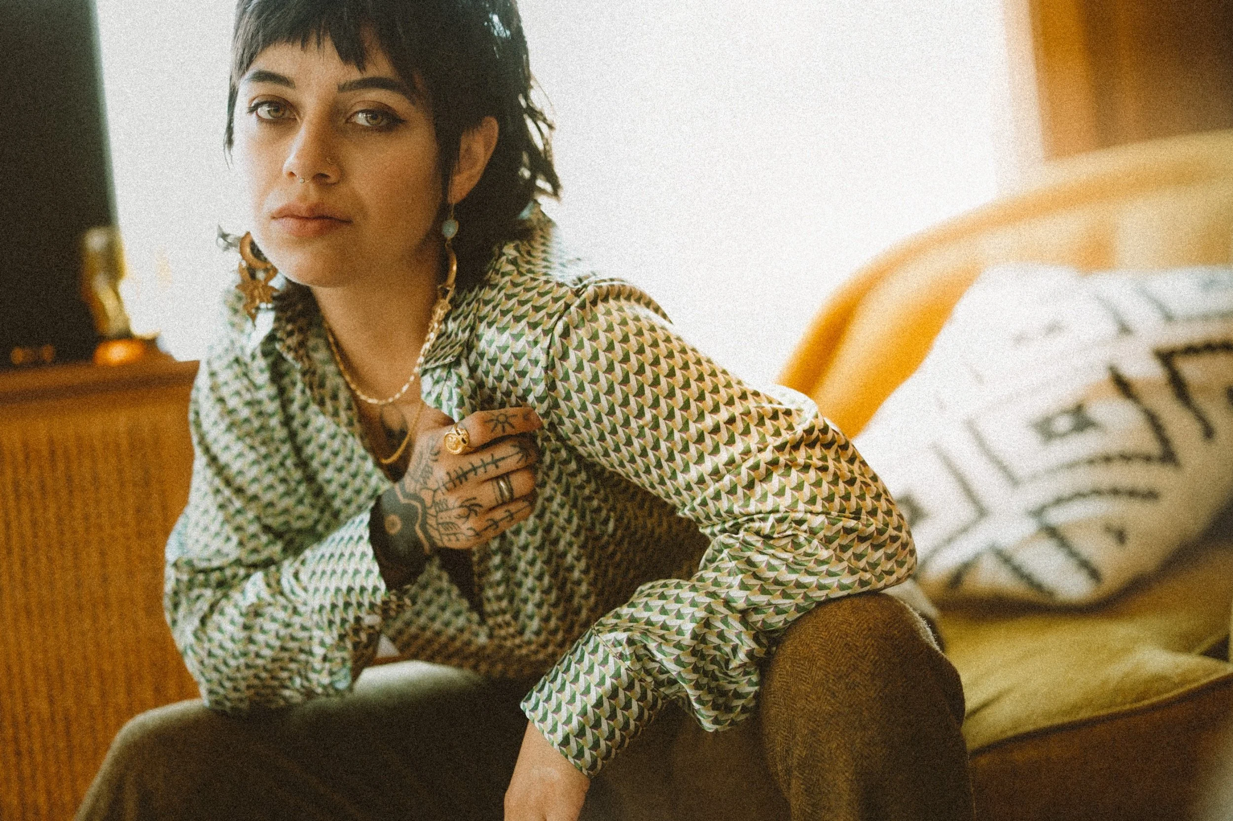 Person with a patterned shirt, tattoos on hands, sitting on a chair.