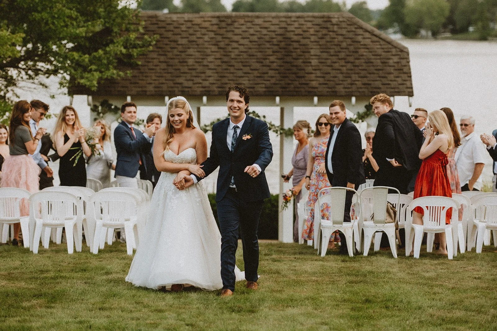 Bride and groom walking down the aisle outdoors after wedding ceremony, surrounded by cheering guests seated on white chairs in a garden setting.