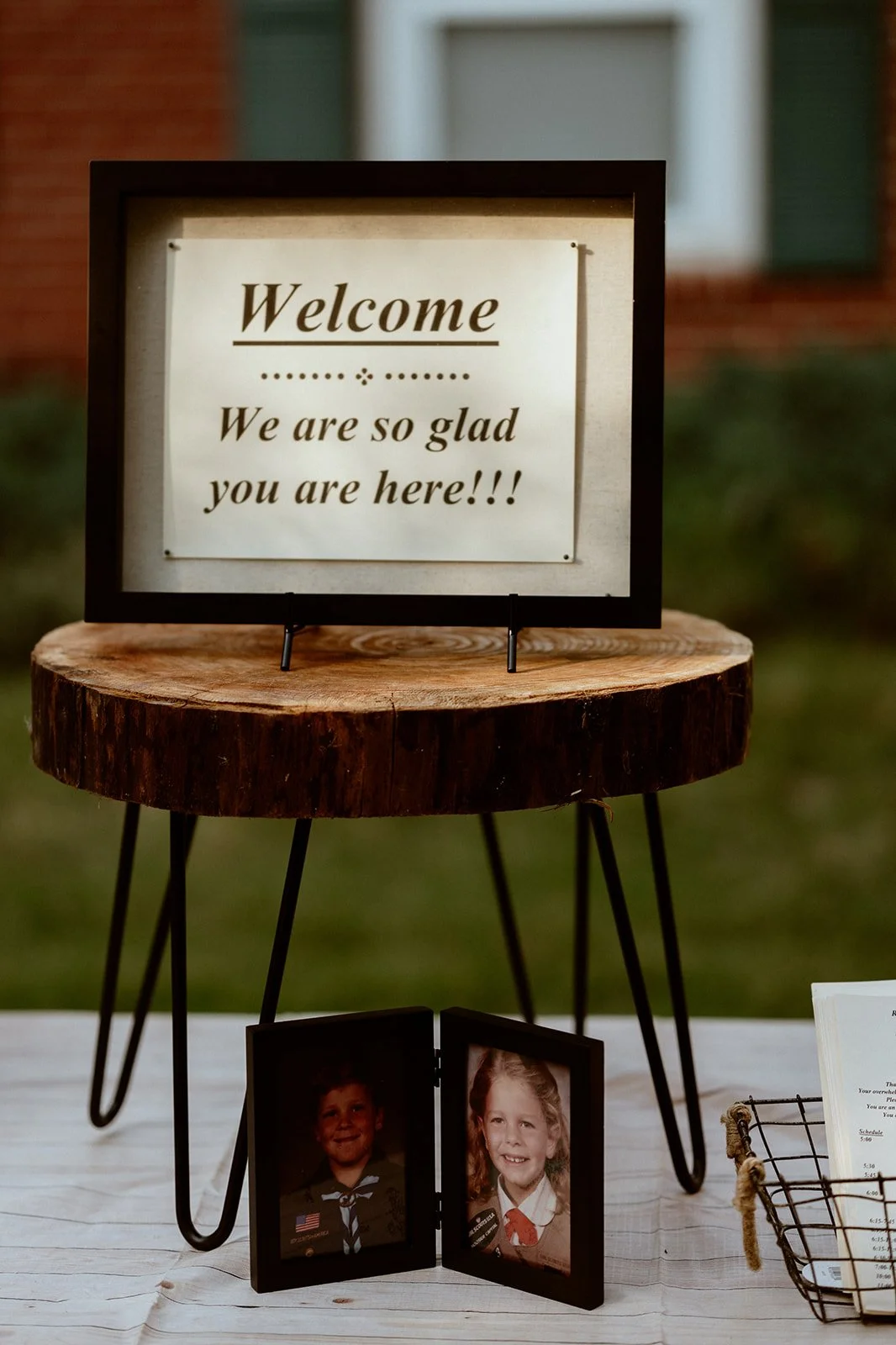 A wooden table with a "Welcome" sign that reads "We are so glad you are here!!!". Below the sign is a small photo frame displaying two childhood portraits. A book or pamphlet and a wire basket are partially visible to the side.