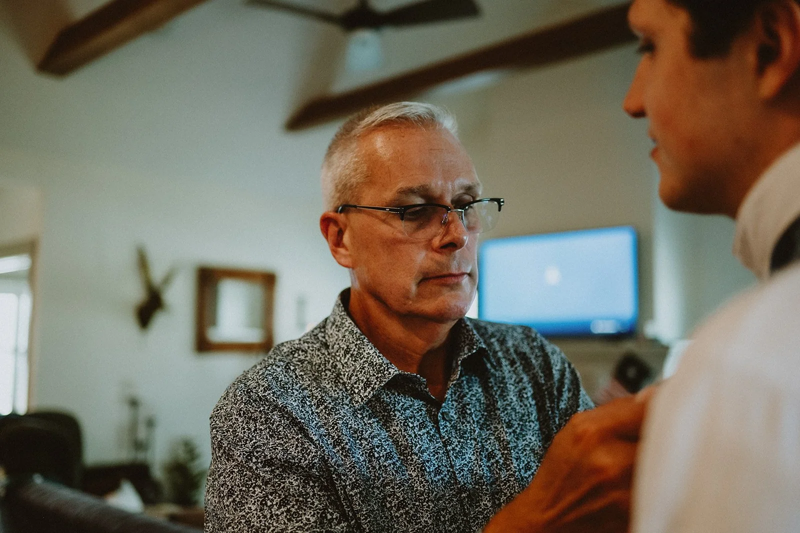 An older man in glasses and a patterned shirt adjusts the tie of another person in a home with a ceiling fan, a framed mirror, and a TV in the background.