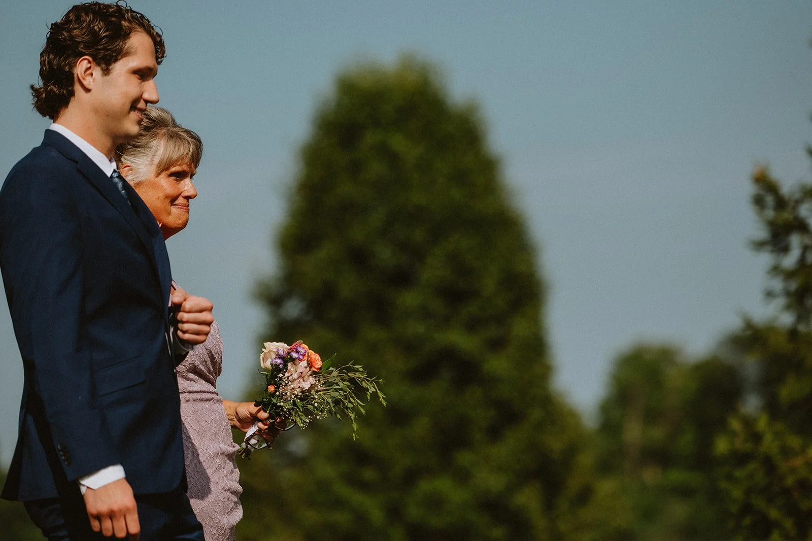 A man in a blue suit walks with a woman holding a bouquet of flowers outdoors.