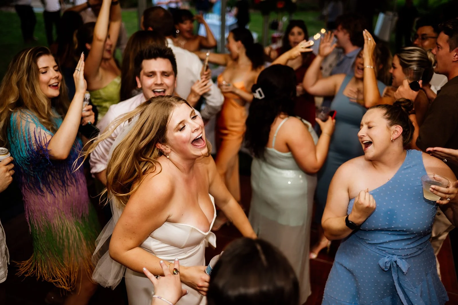 Group of people joyfully dancing at a party, wearing colorful attire and holding drinks.