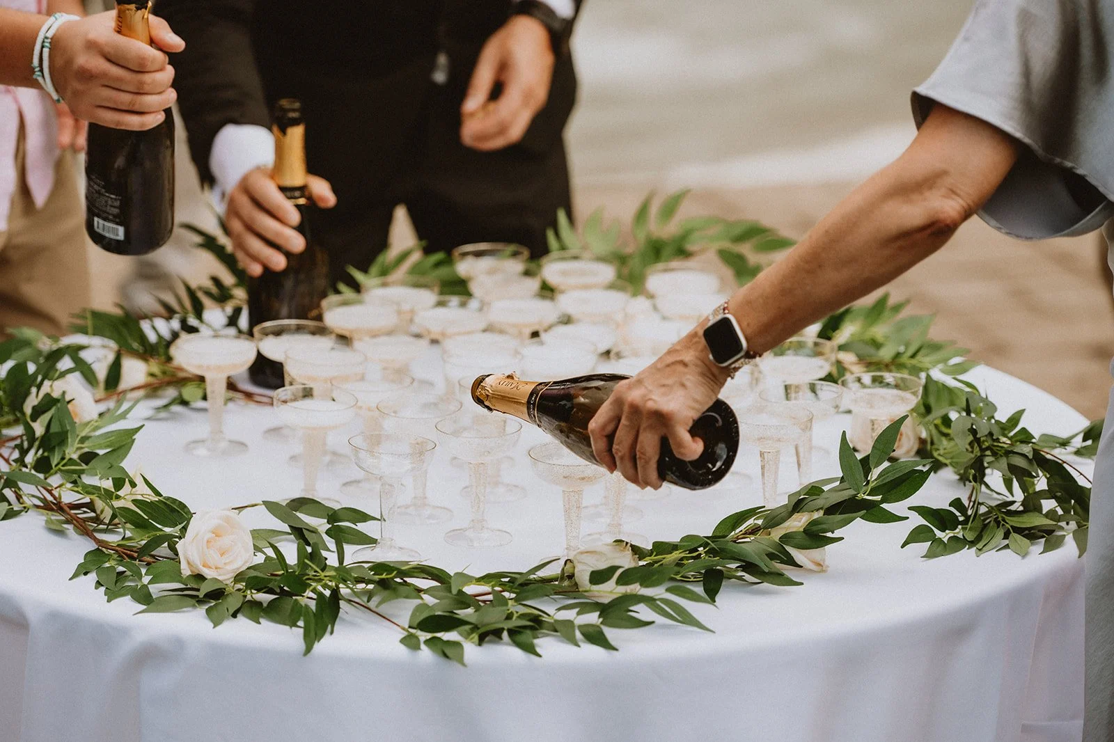 People pouring champagne into glasses on a table decorated with greenery and flowers.