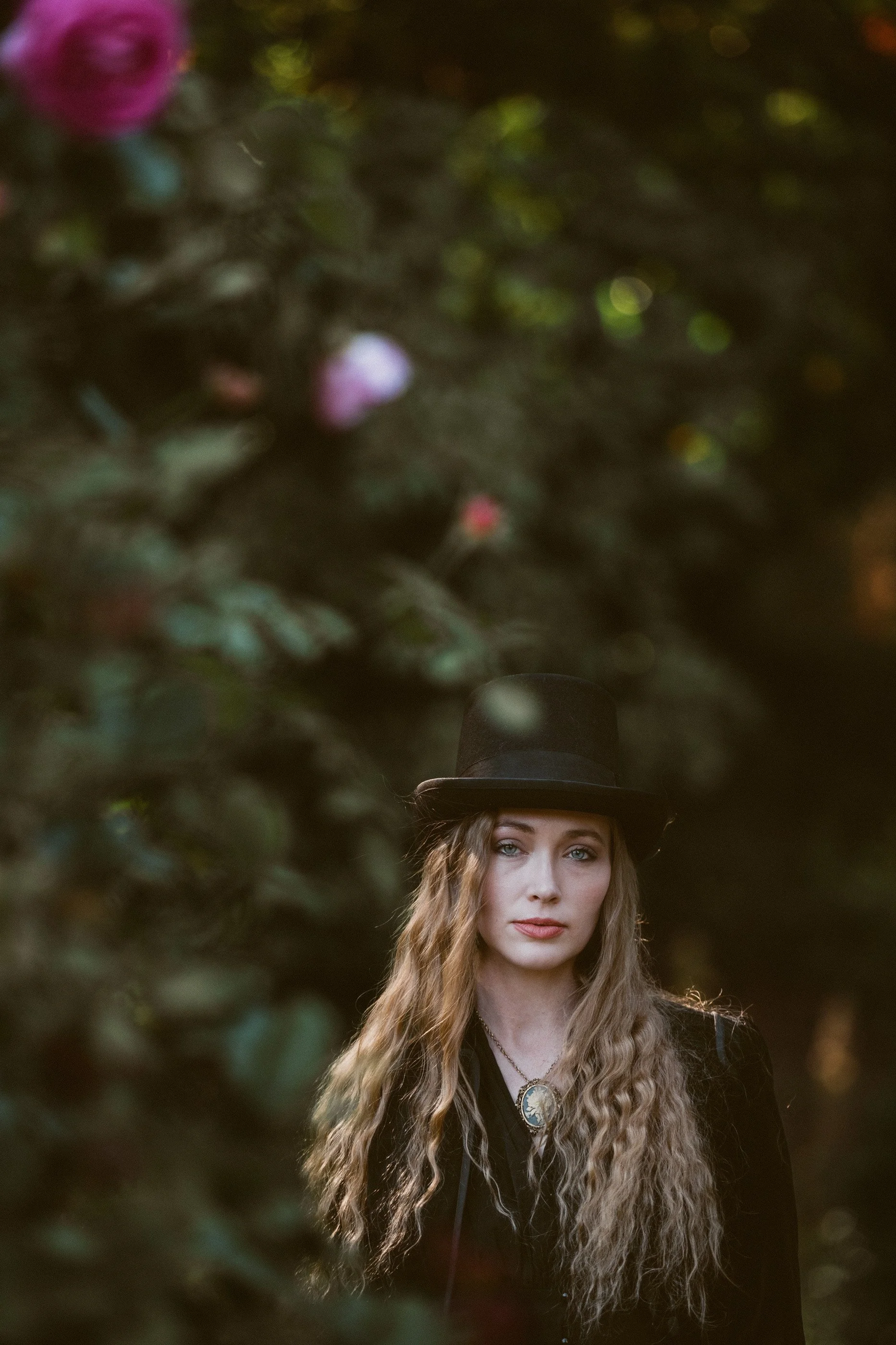 Woman with long wavy hair wearing a black top hat and vintage necklace, standing in front of blurred greenery with roses.