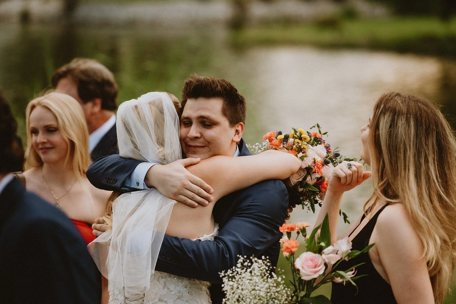 A groom in a navy suit hugging a bride in a white dress with a veil, holding a colorful bouquet, near a lake, with wedding guests nearby.