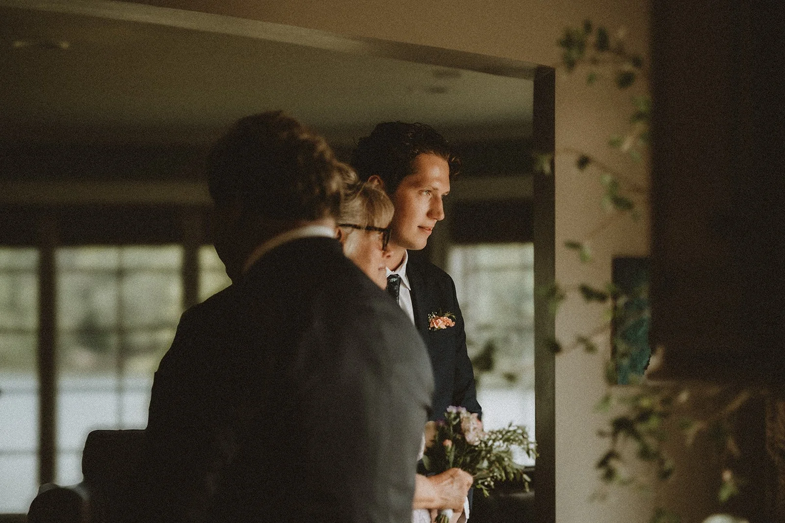 Man in a suit holding flowers, standing with another person indoors, soft lighting.