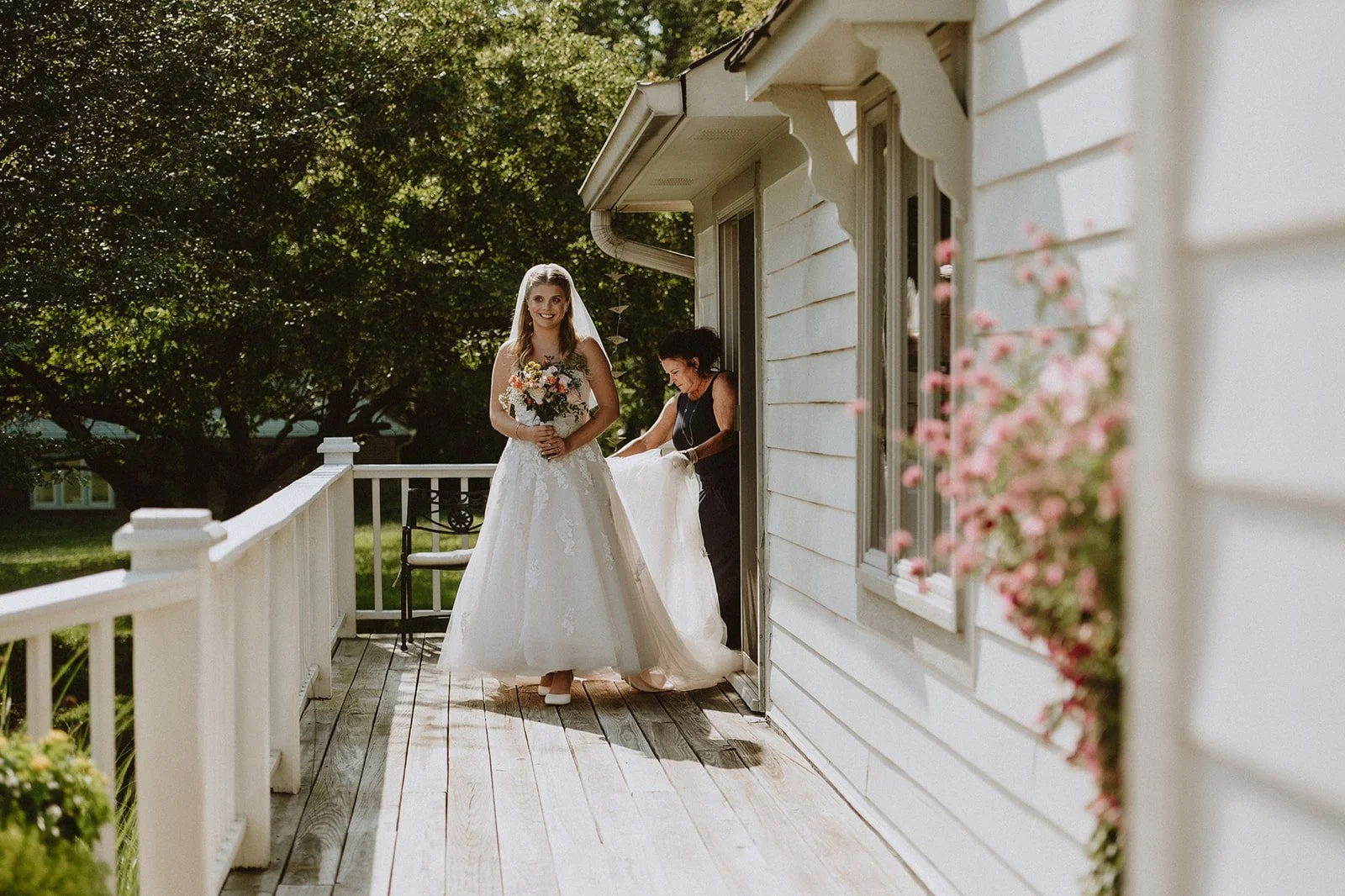 Bride in white dress holding bouquet on porch with woman adjusting dress, flowers and greenery surrounding them.