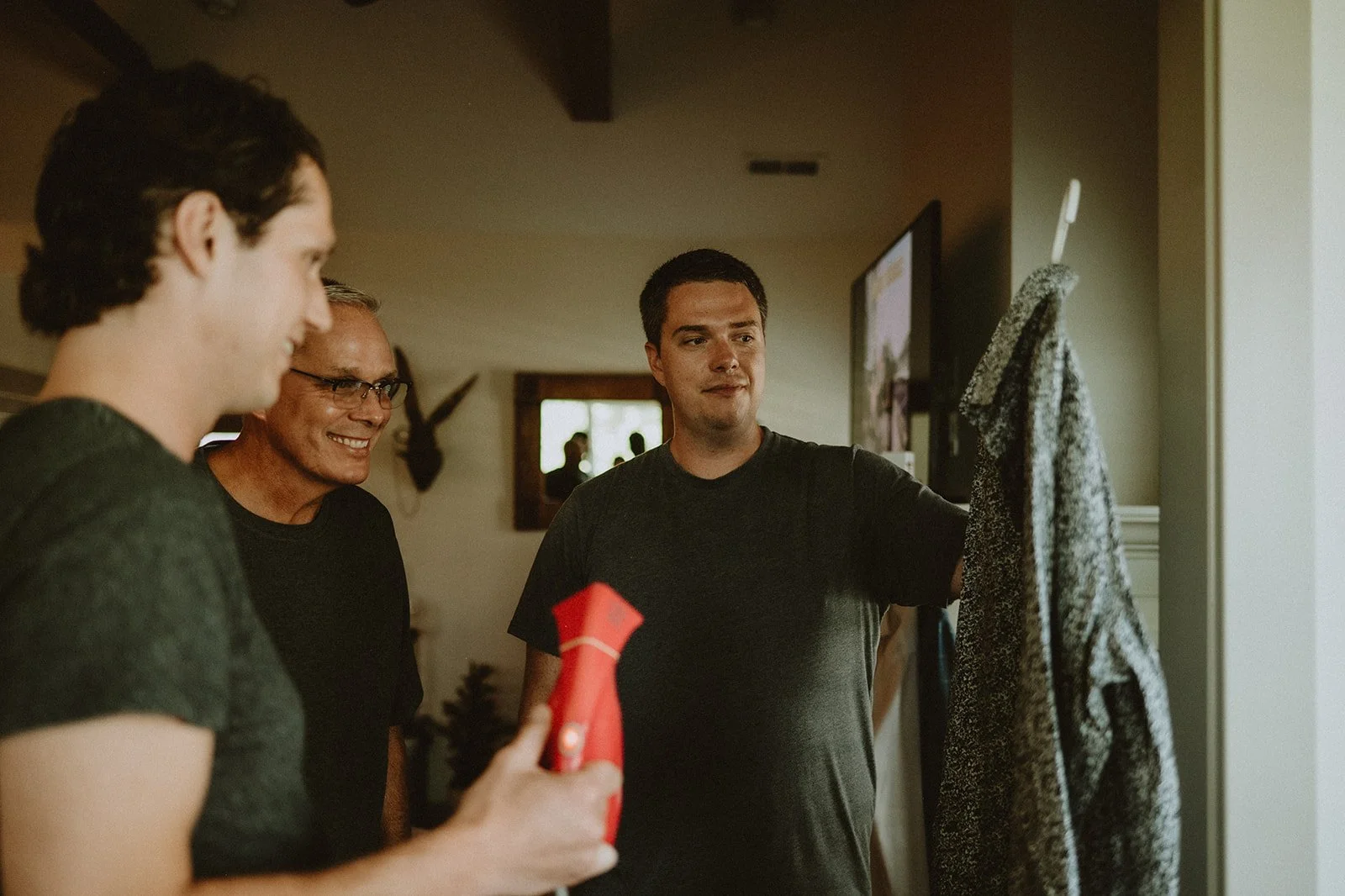 Three men in a room, one holding a garment steamer and another holding a coat on a hanger.