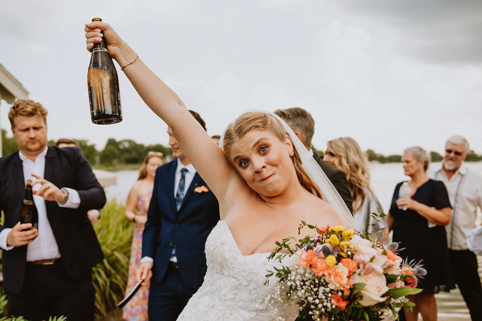 Bride holding a champagne bottle and bouquet, surrounded by wedding guests.