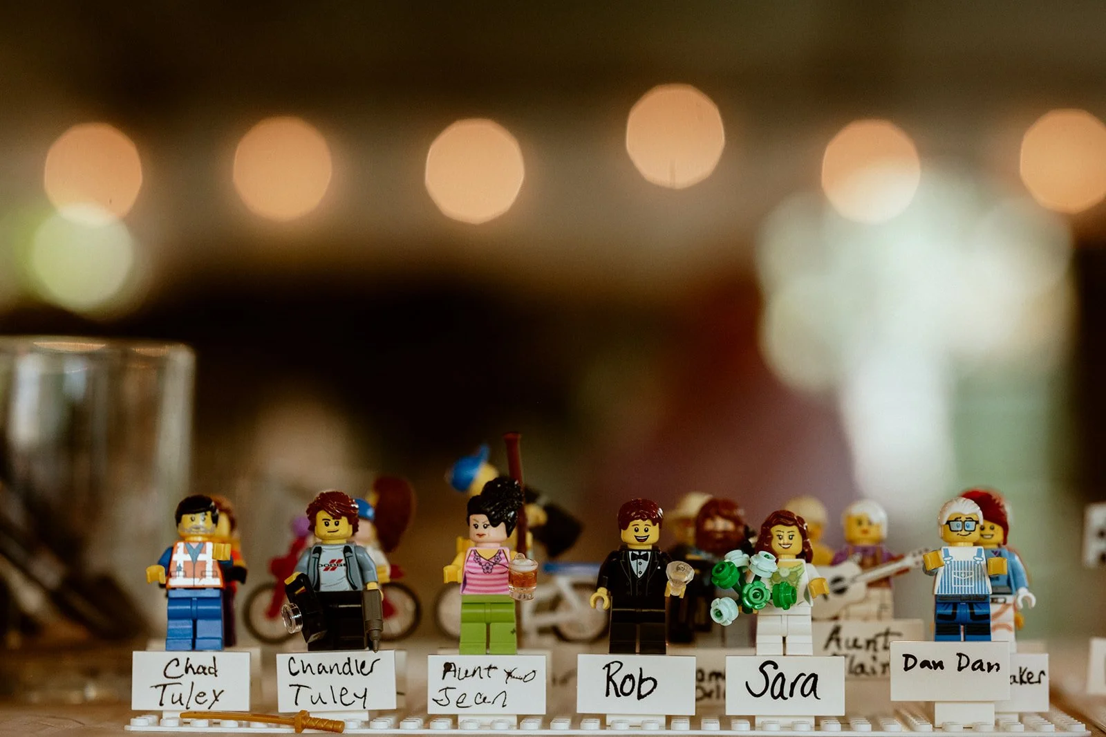 Close-up of Lego minifigures with name cards, including figures in various outfits like construction gear, casual clothing, and formal wear, against a blurred background with lights.