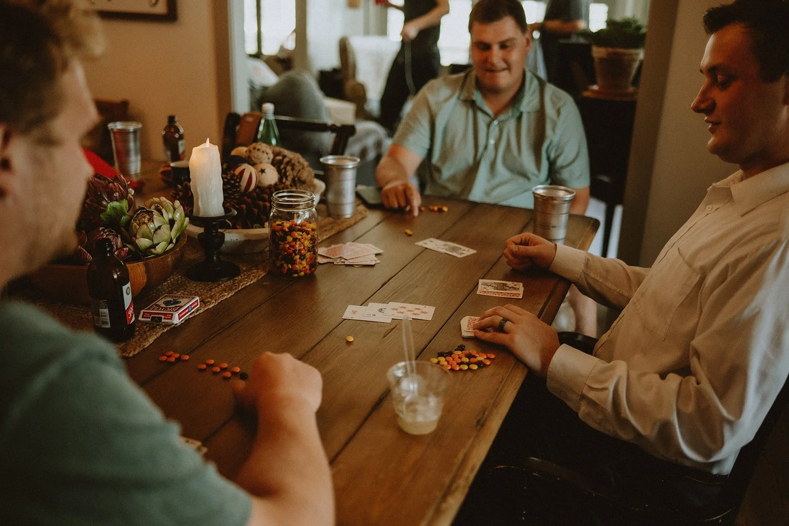 Three men sitting around a table playing cards with candies used as poker chips; a candle, jar of candies, and beverage bottles are also on the table.