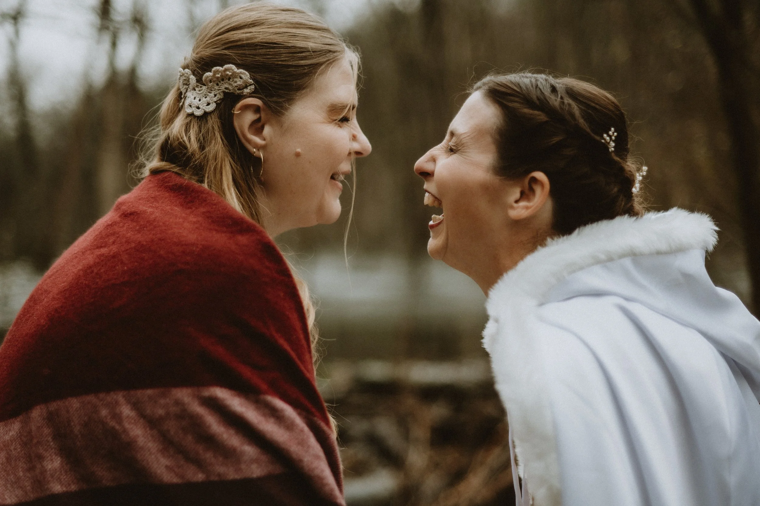 Two women laughing outdoors, one wearing a red shawl and the other a white cape with fur trim, with trees blurred in the background.