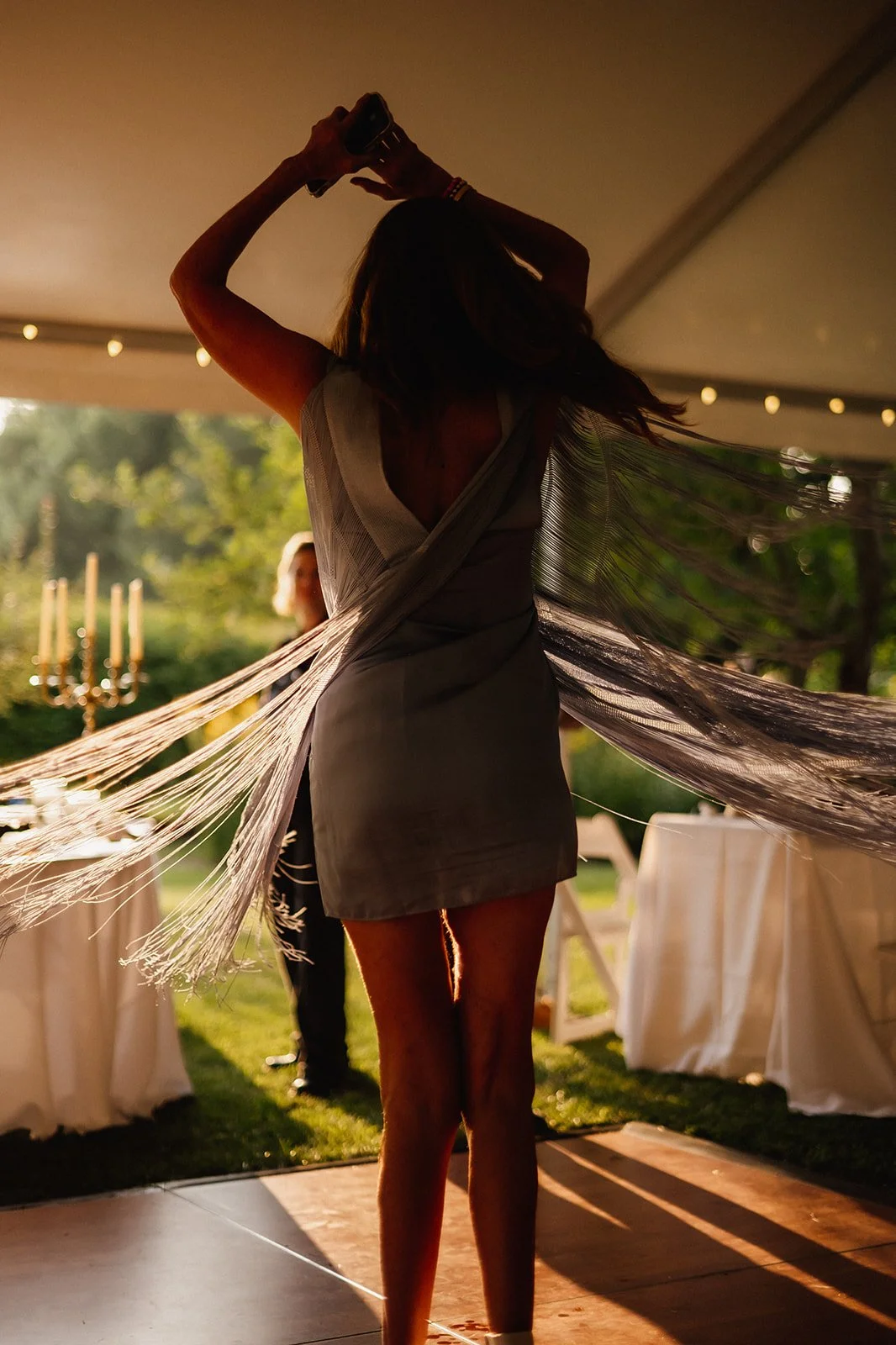 Person dancing at outdoor event, wearing a fringed dress, holding a phone, with white tables and chairs in the background.