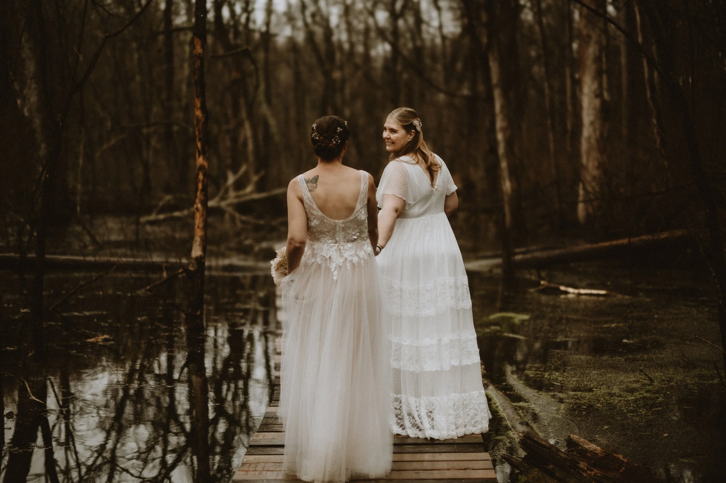 Two women in white dresses standing on a wooden path in a forested, swampy area.