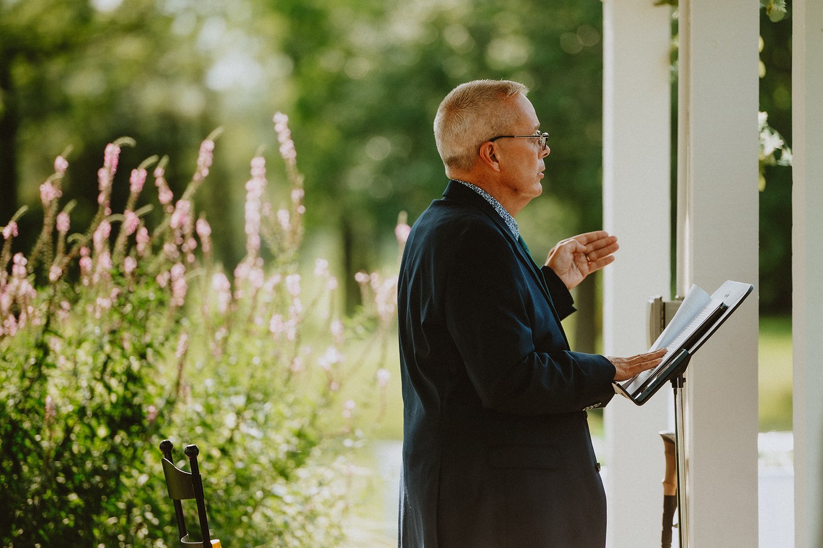 Man in a suit standing outdoors, holding and gesturing towards a book on a stand, with greenery in the background.