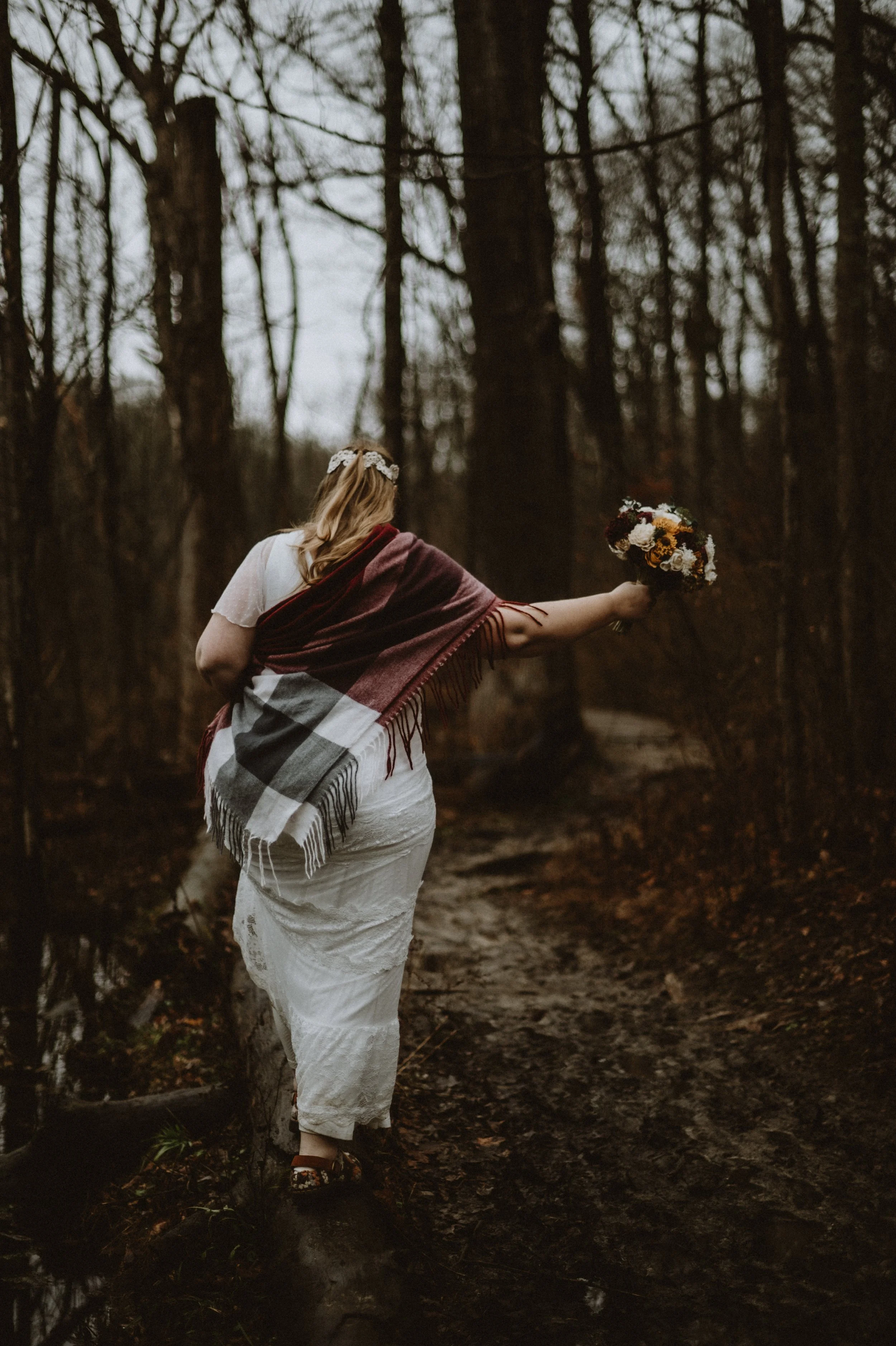 Woman in white dress and shawl walking in a forest with a bouquet