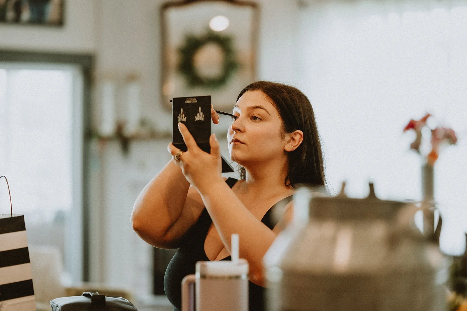 Woman applying makeup using a compact mirror, with a focused expression, in a cozy home setting.