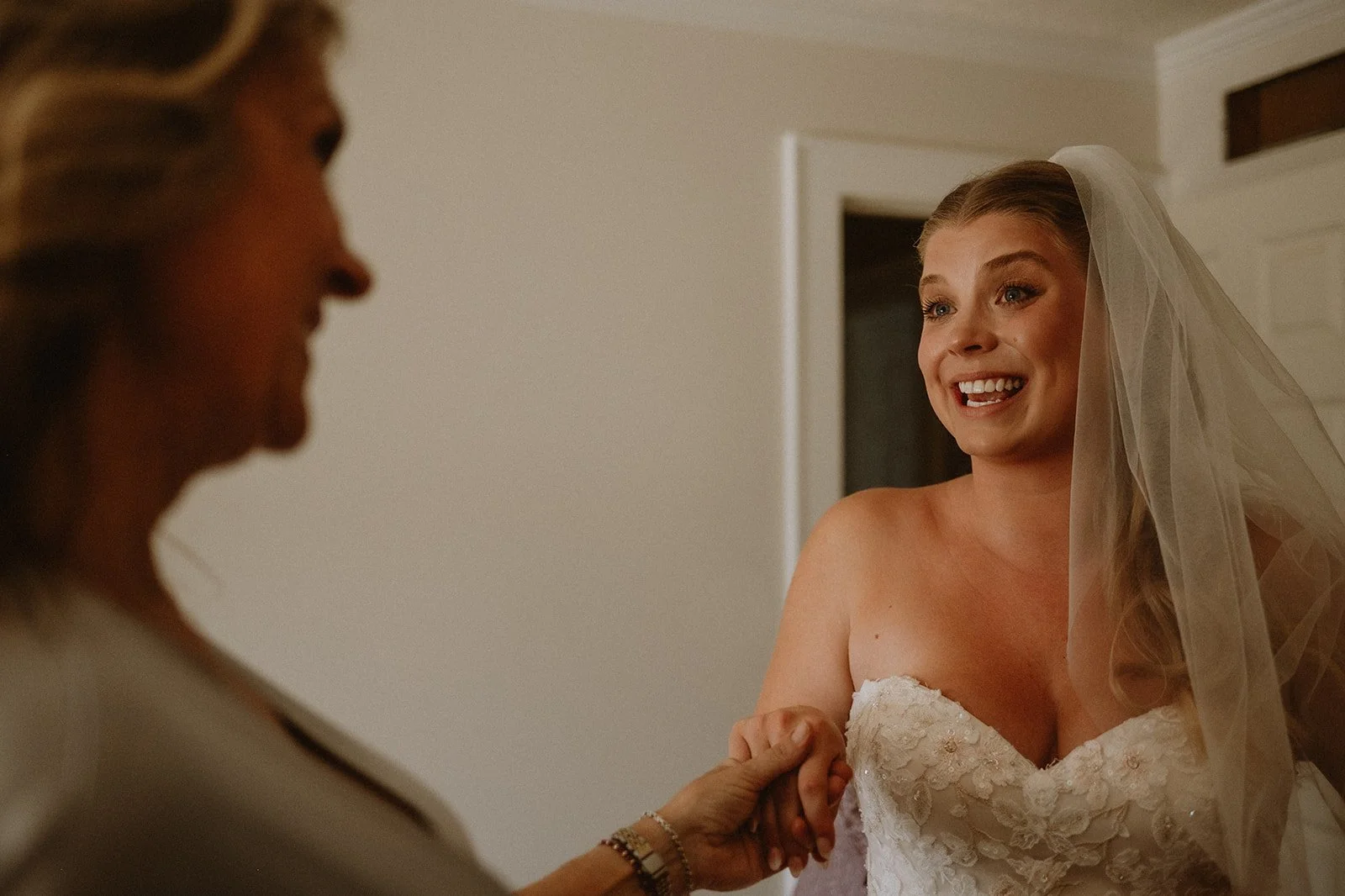 Bride in white wedding dress smiling and holding hands with another person indoors.