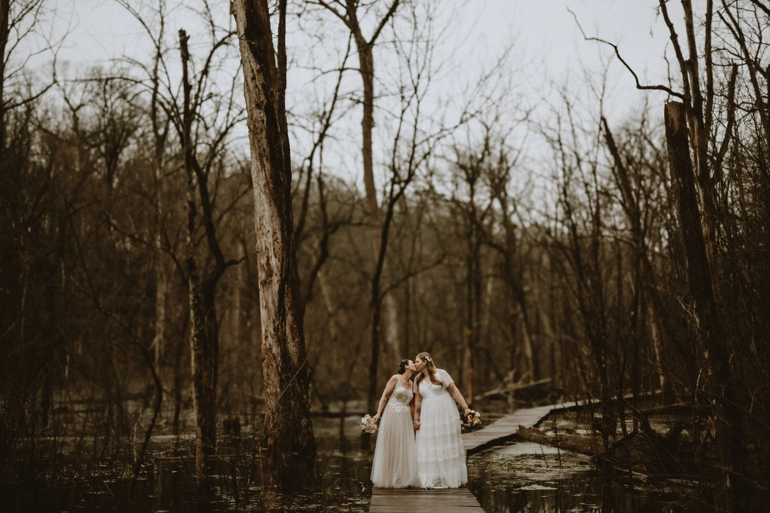 Two brides in wedding dresses kissing on a wooden path in a forest.