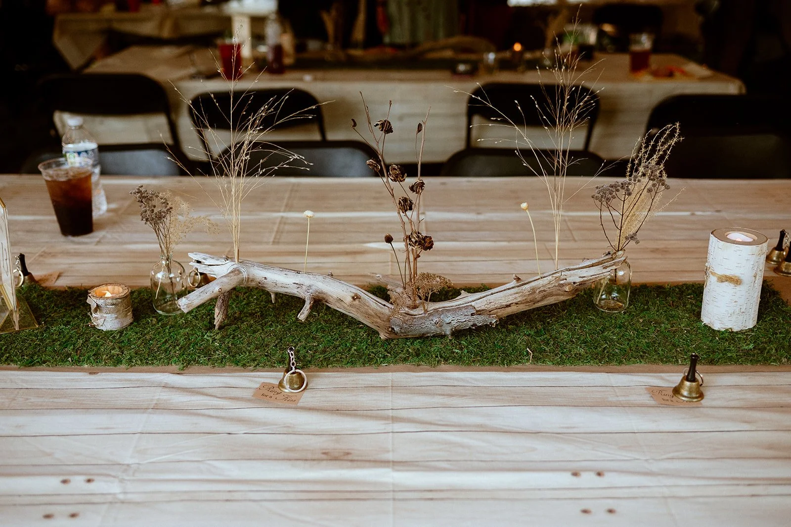 Rustic table centerpiece with driftwood, dried grass, glass vases, candles, and moss on a wooden table with drinks in the background.