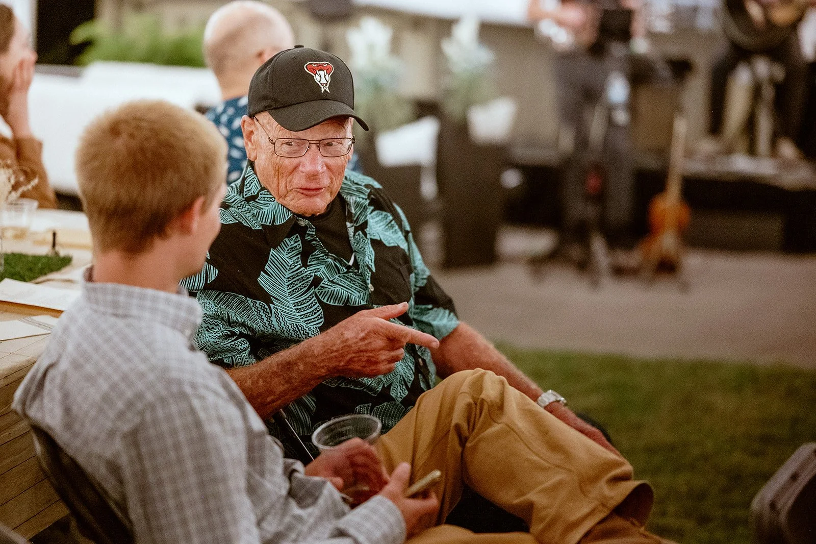 Two men sitting at an outdoor event, engaged in conversation. One is wearing a cap and a patterned shirt, pointing with his finger. The other is holding a drink. People are blurred in the background.
