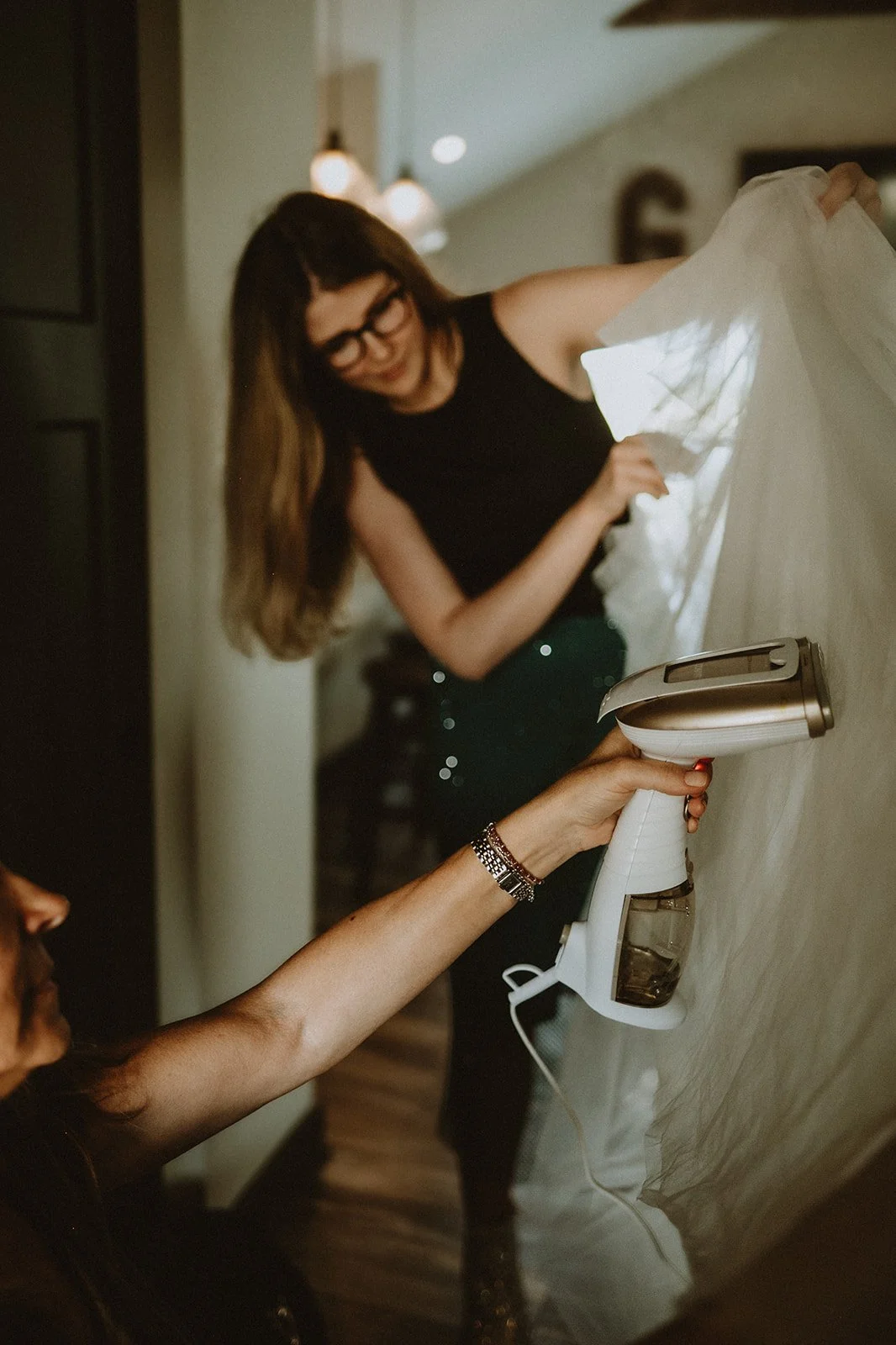 Person using a handheld steamer on a garment while another person holds the fabric, inside a dimly lit room with pendant lights and wooden floor.