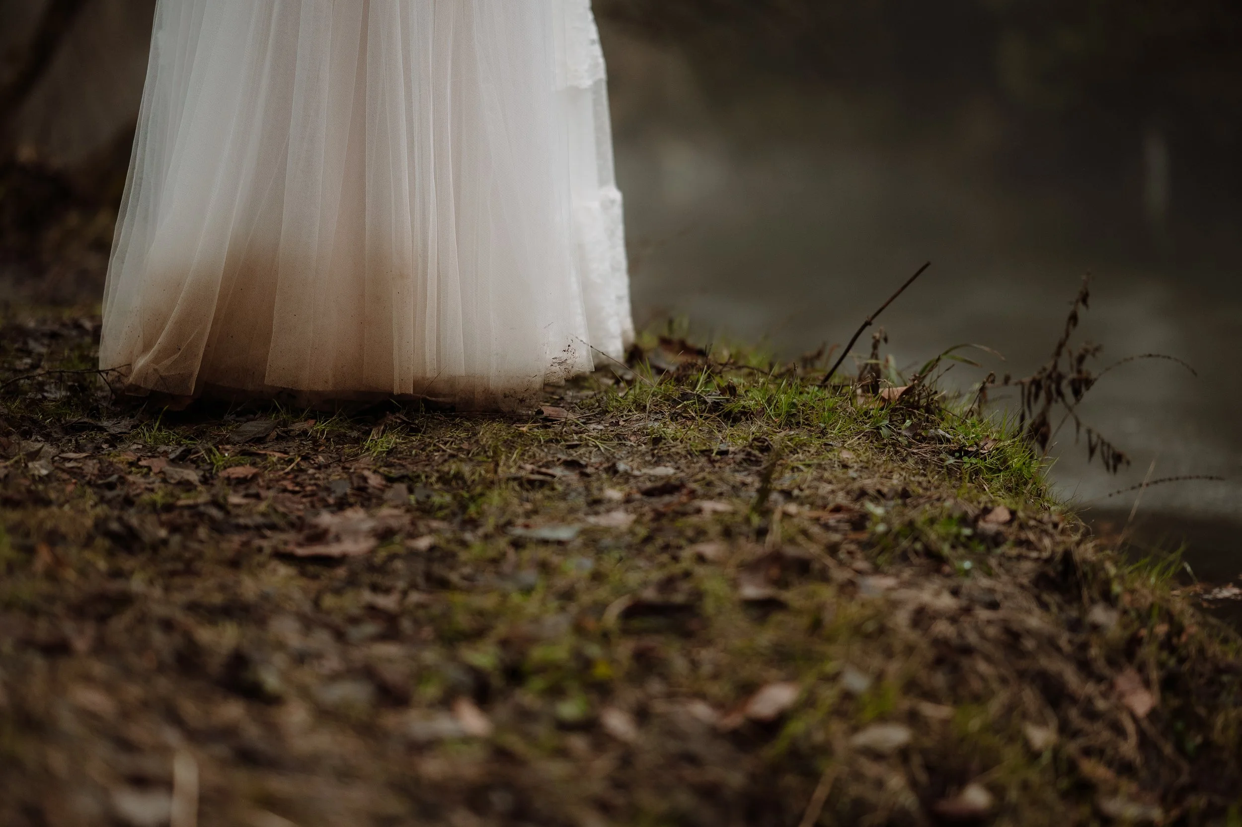 Close-up of a bridal gown's train dragging on a muddy, grassy ground near a body of water.