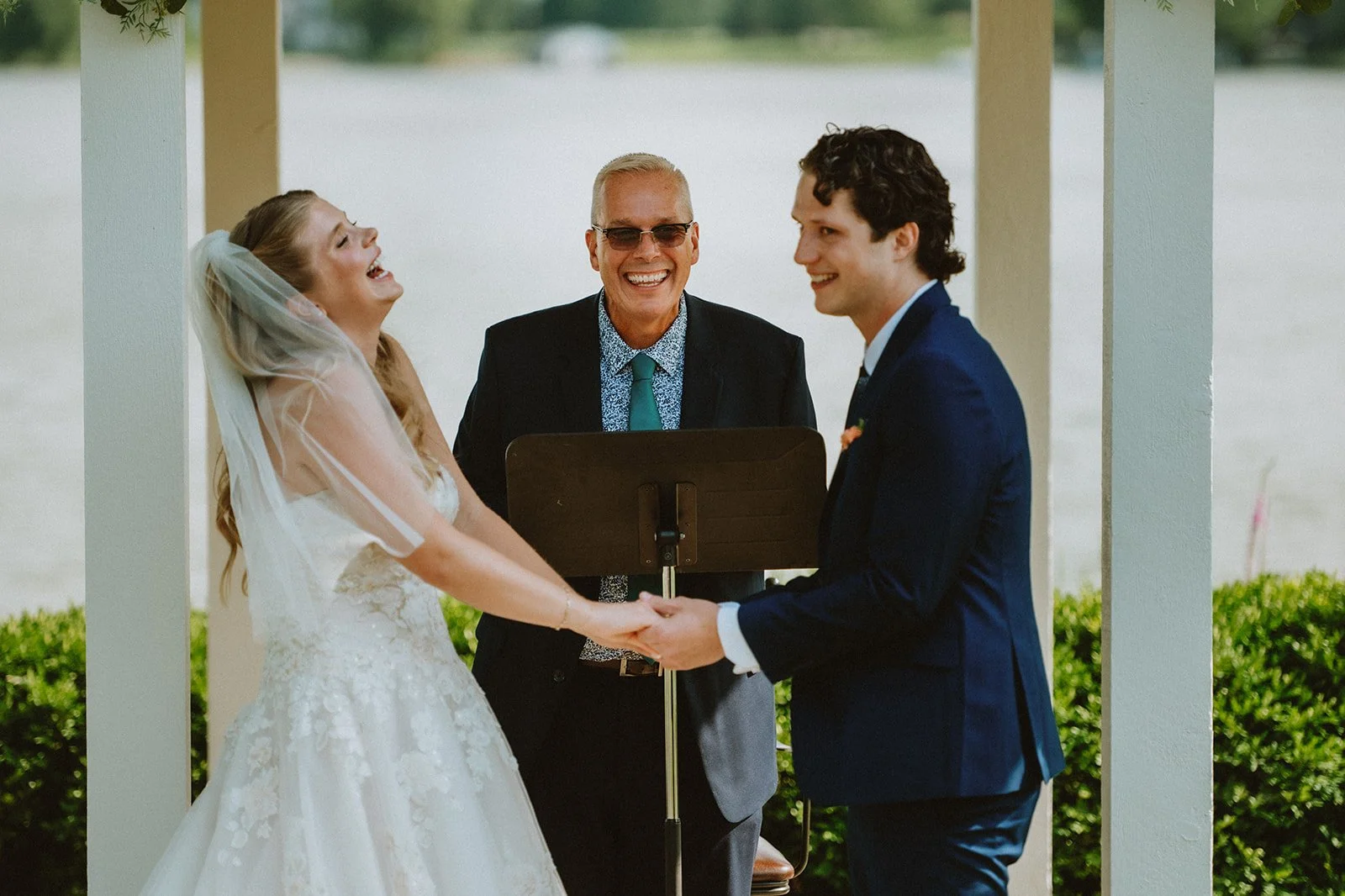 Bride and groom holding hands and laughing with officiant during outdoor wedding ceremony.