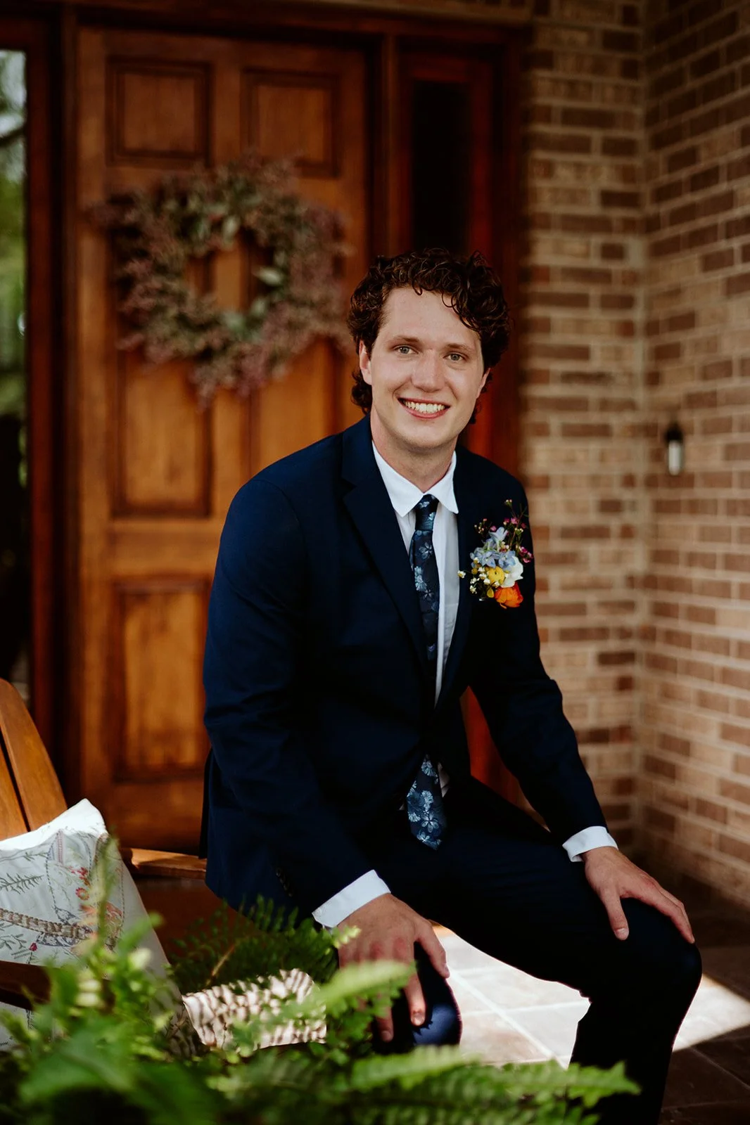 Smiling man in a blue suit sitting on a porch with a brick wall and wooden door in the background.