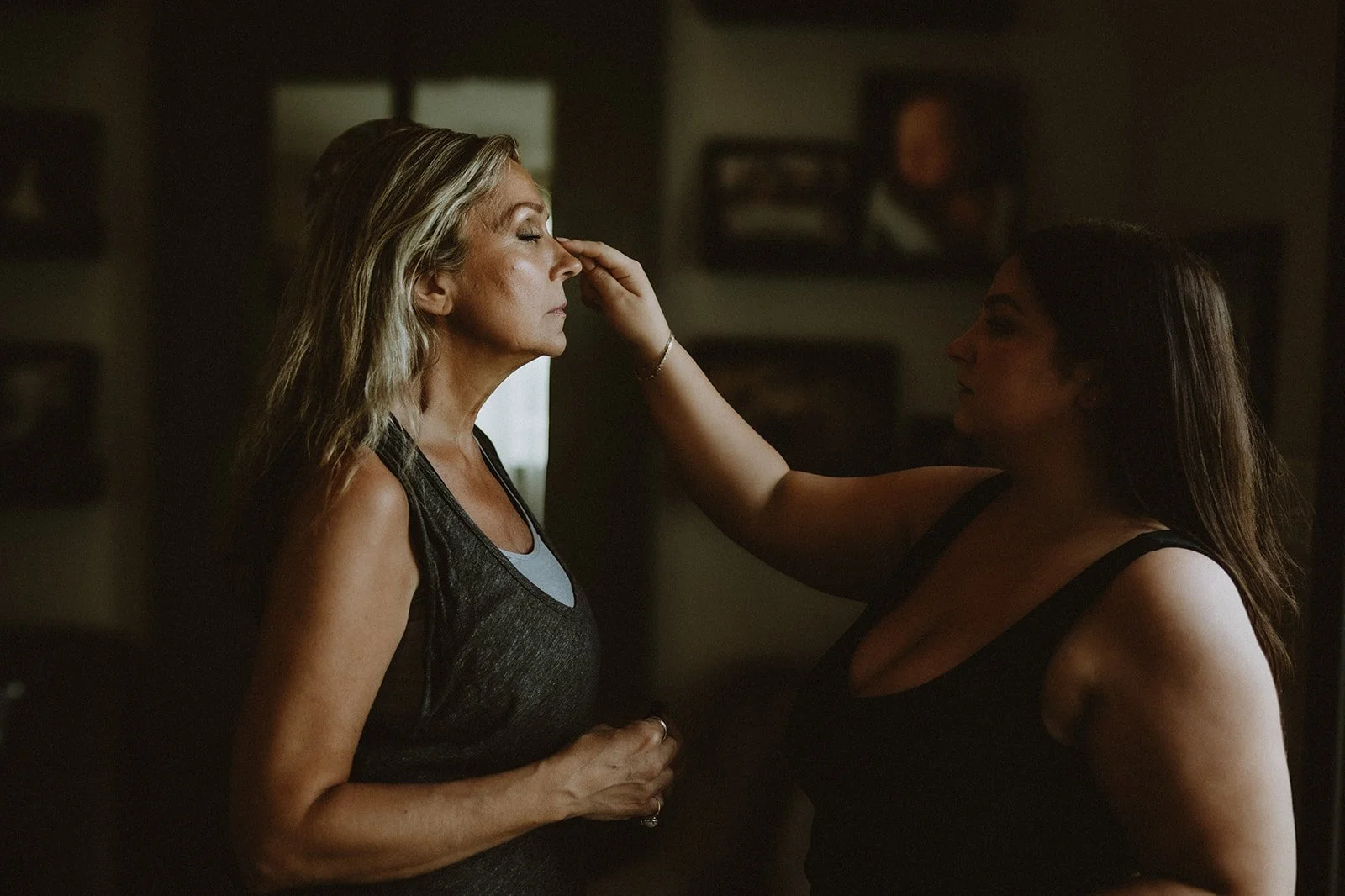 Woman applying makeup to another woman's face in a dimly lit room.