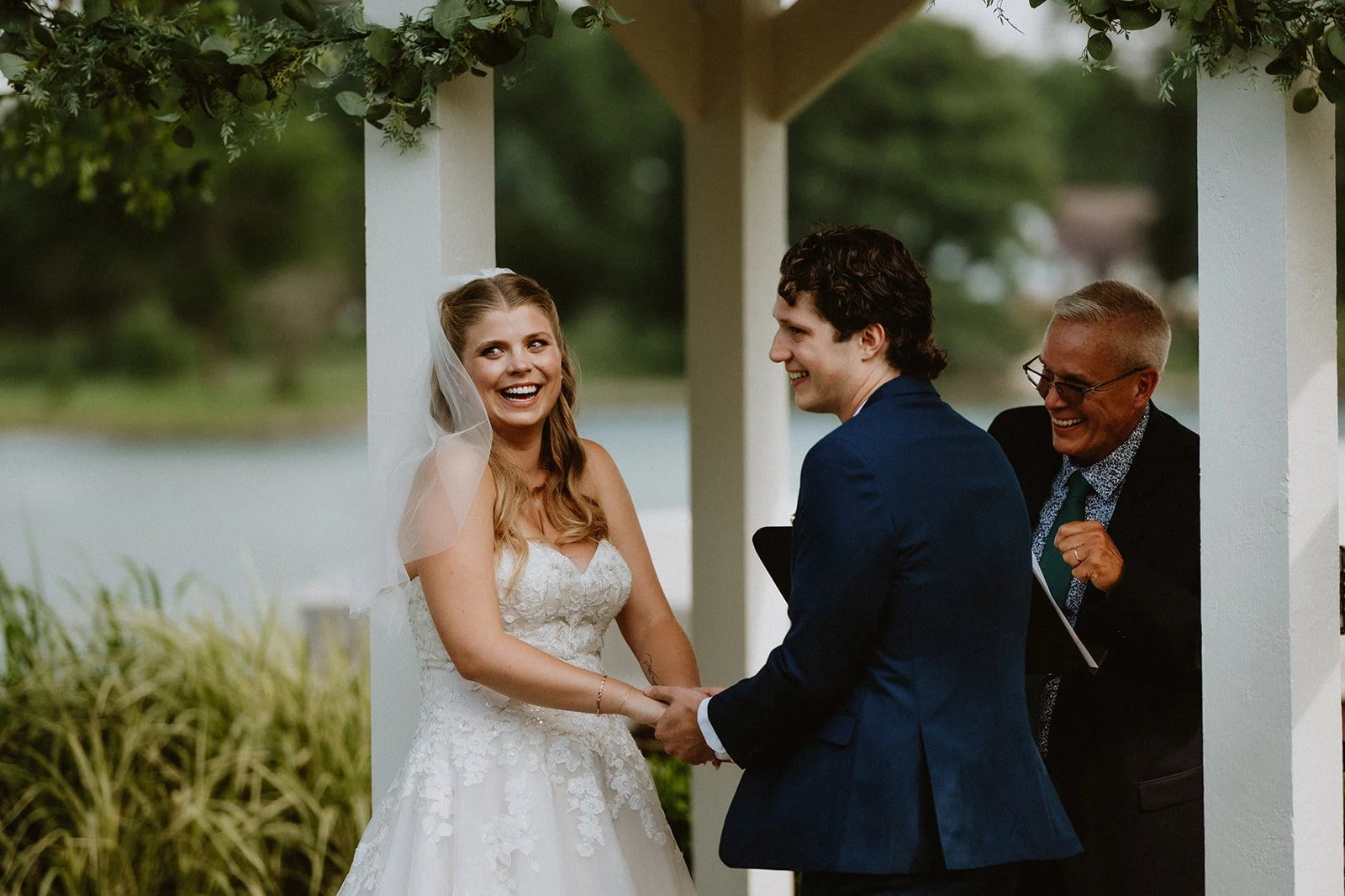Bride and groom smiling during outdoor wedding ceremony with officiant laughing nearby.