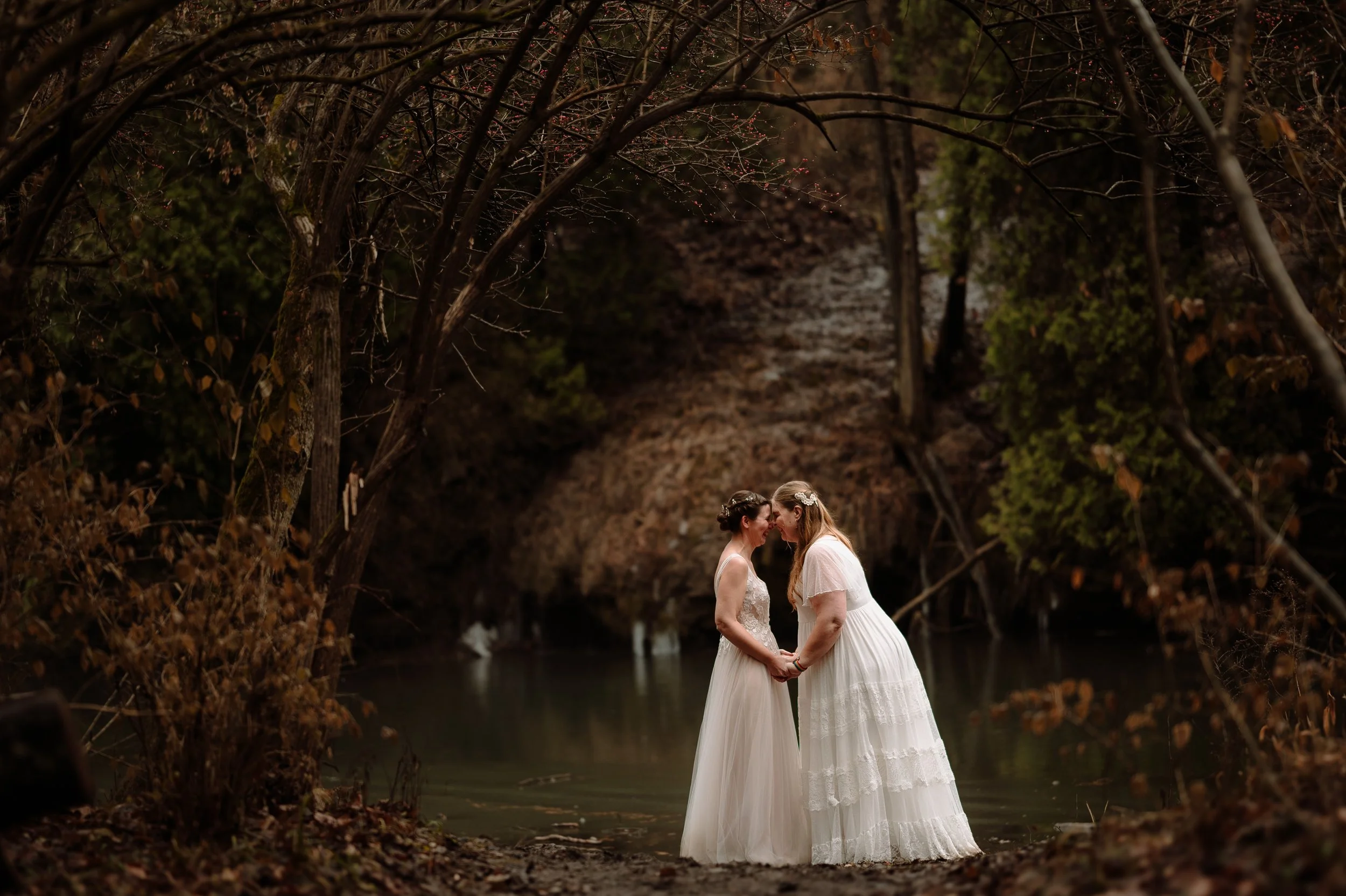 Two women in wedding dresses standing by a lake surrounded by trees, holding hands and leaning foreheads together.