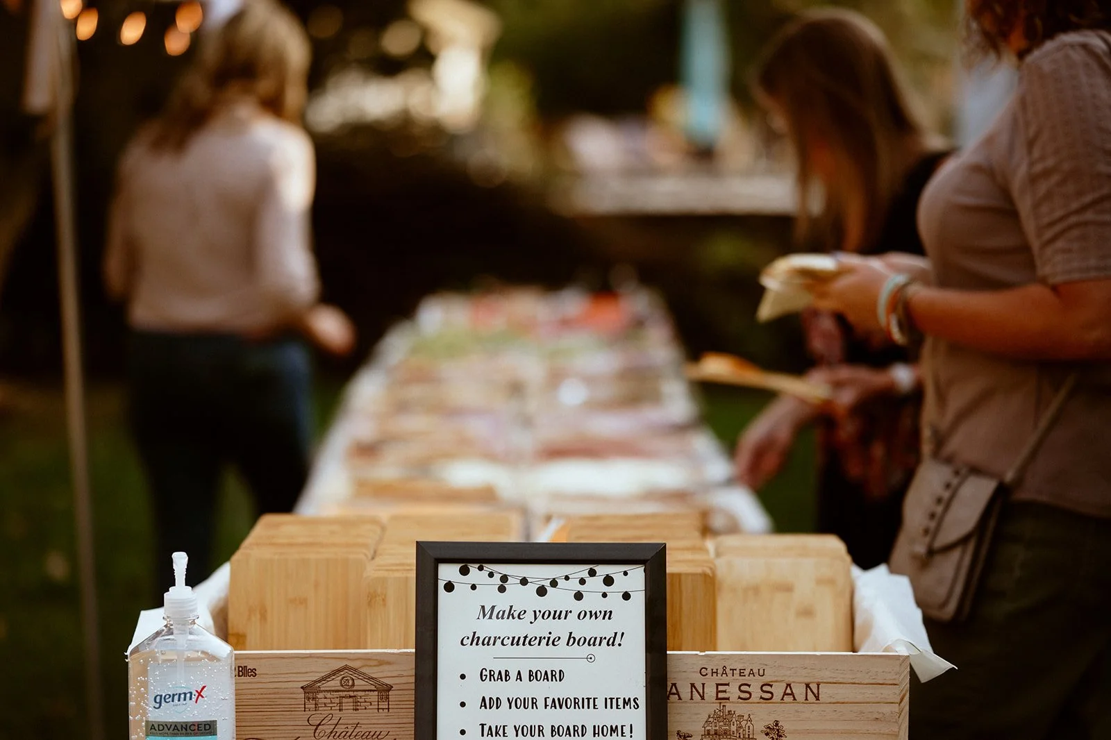 Guests creating charcuterie boards at an outdoor event with a sign instructing on how to make your own board, next to hand sanitizer and wooden boards.