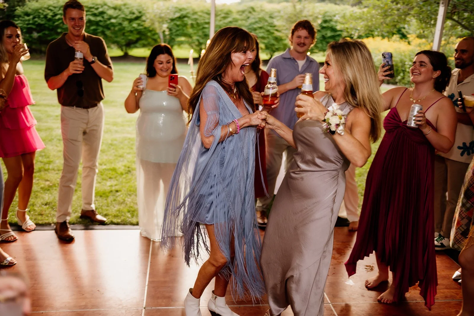 Group of people dancing and celebrating at a party or event outdoors, with women in colorful dresses holding drinks. They appear joyful and engaged, set against a background of greenery.