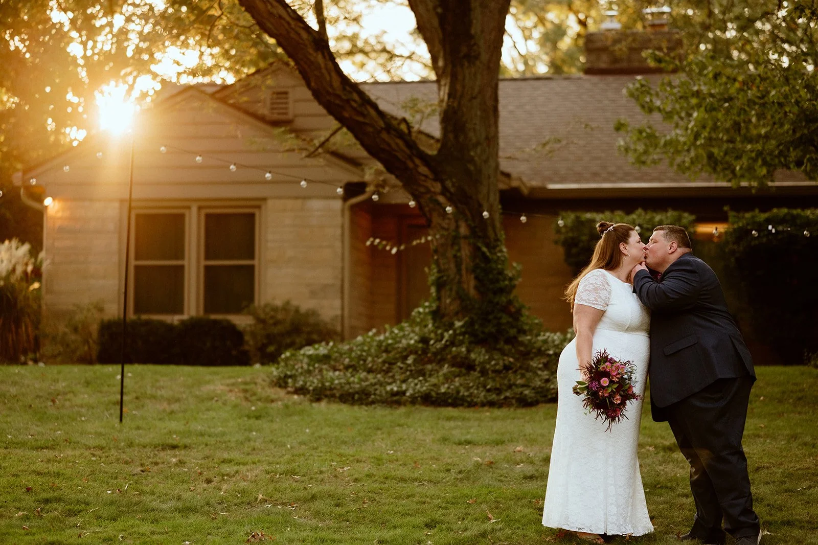 A couple kissing in front of a house at sunset, with the woman in a white dress holding a bouquet, and the man in a dark suit. A large tree is in the background.