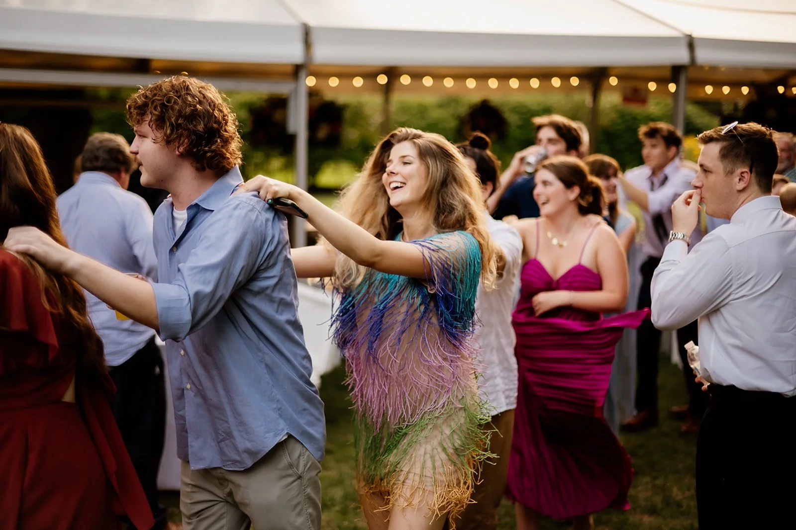 People dancing in a conga line at an outdoor party under a tent with string lights.