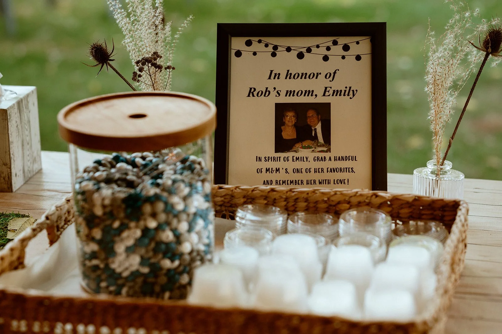 Memorial display for Rob's mom, Emily, featuring a framed photo and message inviting guests to take M&M's, set on a wooden table with dried flowers and a basket of small jars.