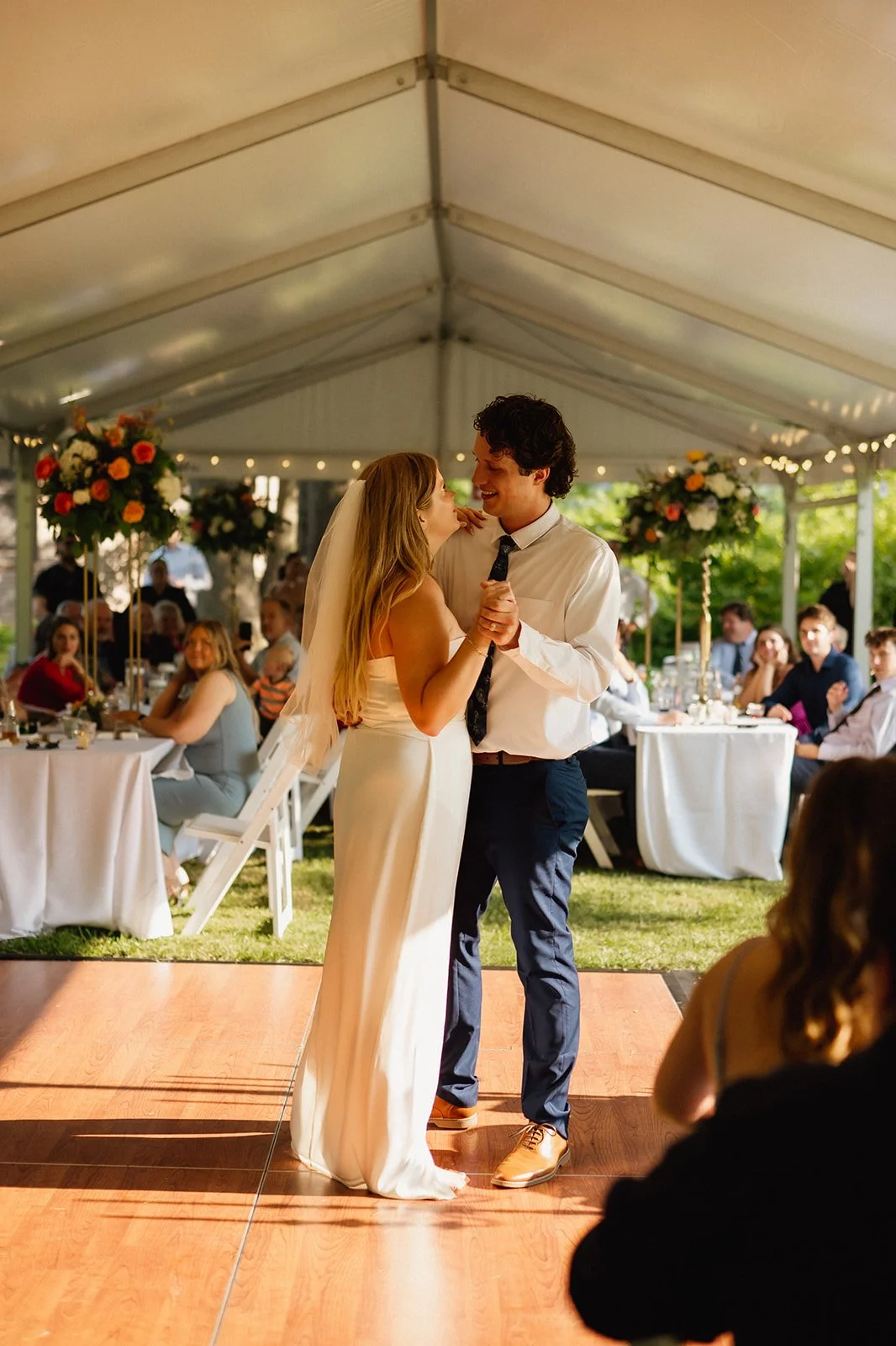Bride and groom dancing at wedding reception under a tent, surrounded by guests seated at tables with floral centerpieces.