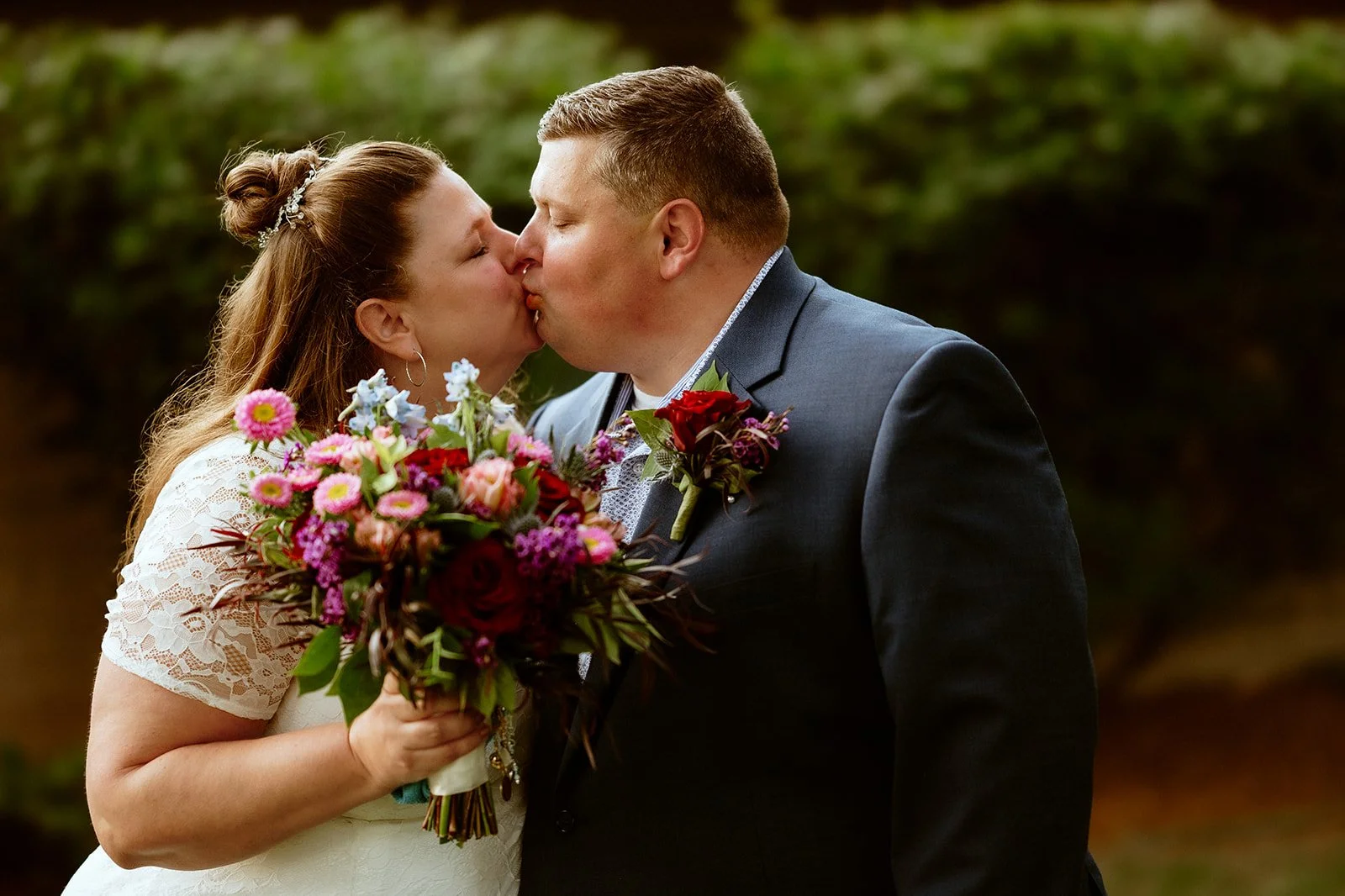 A bride and groom kissing, with the bride holding a colorful bouquet, outdoors.