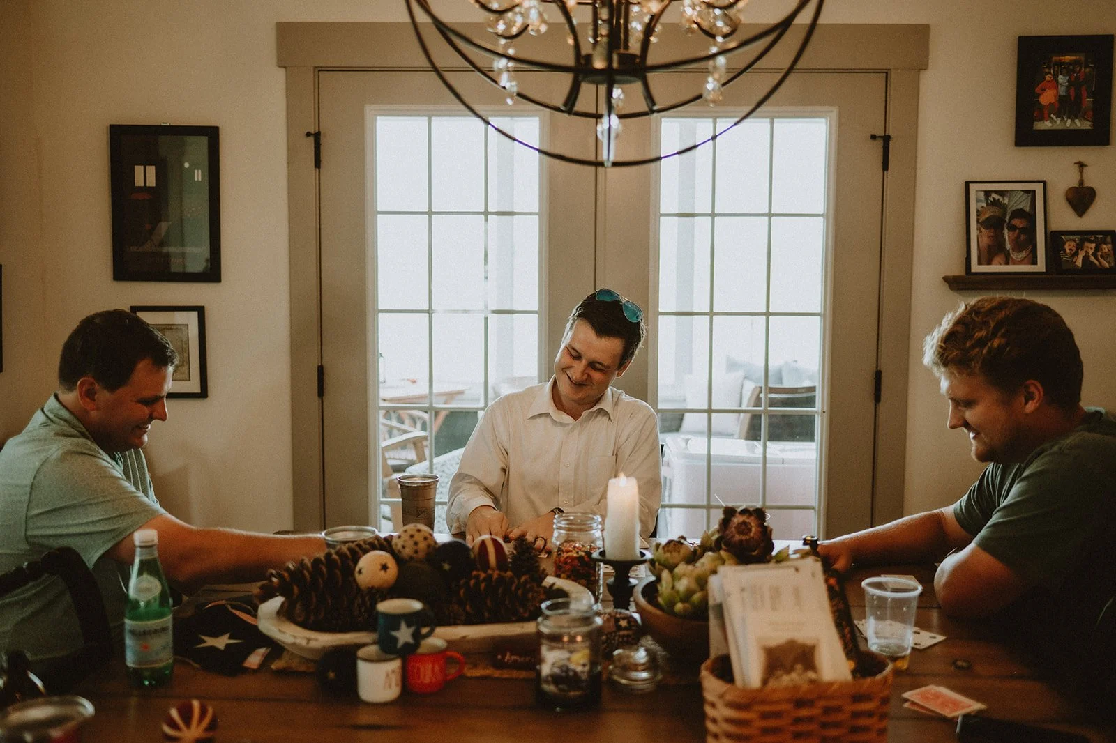Three men sitting around a dining table playing a game, surrounded by decorative items and drinks, with a chandelier overhead.